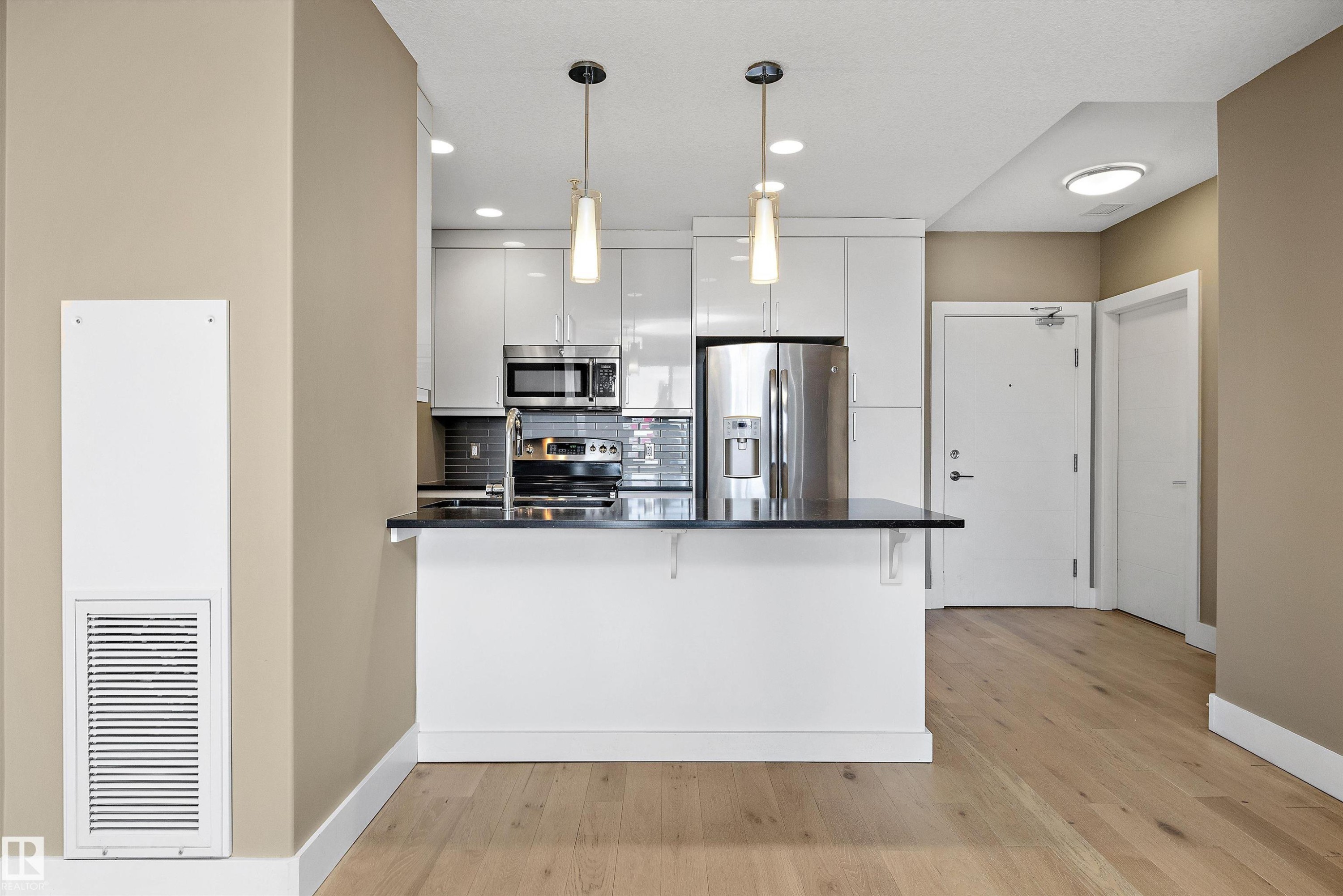 Contemporary kitchen featuring high-gloss white cabinetry, stainless steel appliances, dark countertop, and pendant lighting over a breakfast bar - 307 11080 Ellerslie Rd Sw, Edmonton, AB