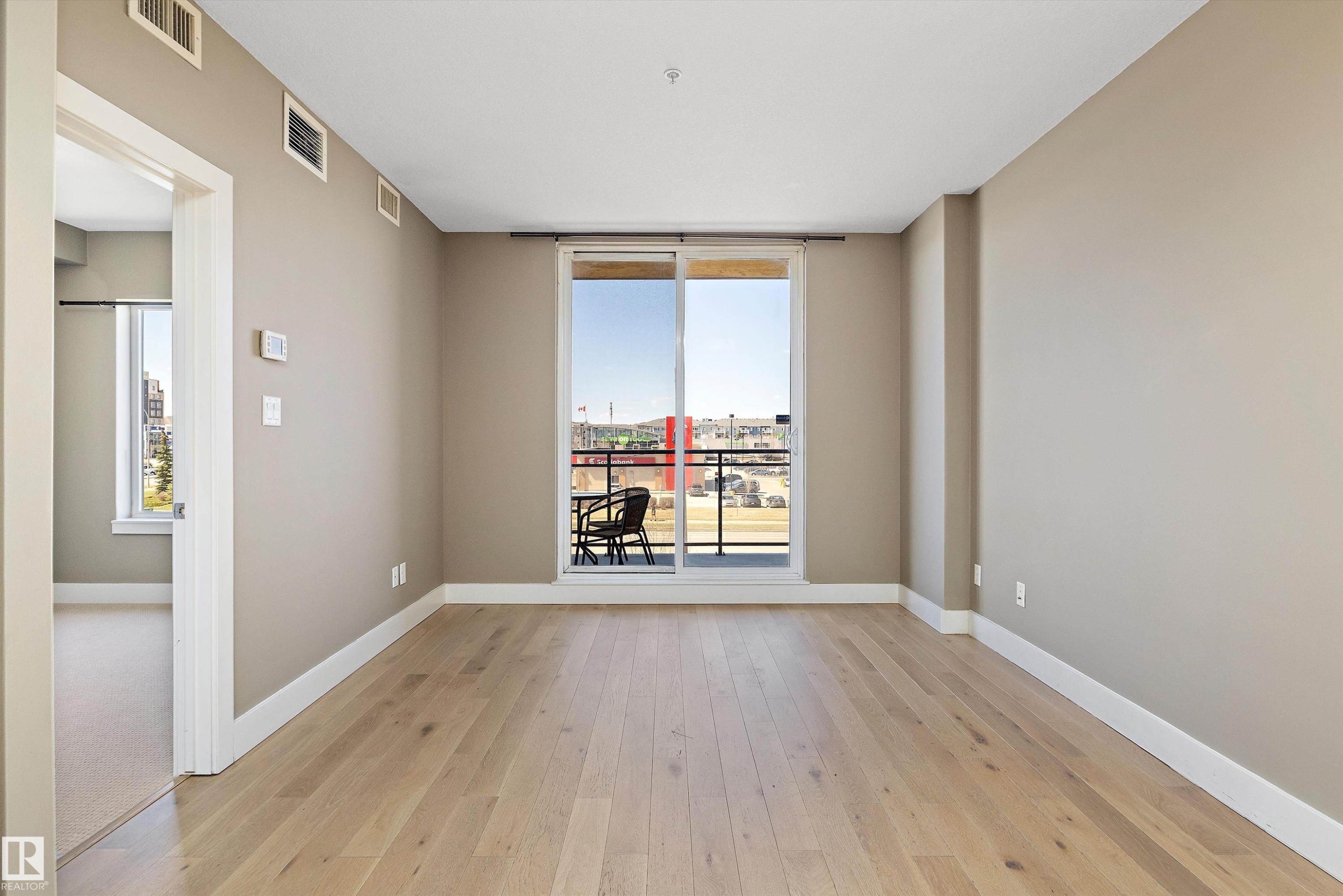 Spacious room featuring wood-finish flooring, light-toned walls, and a sliding glass door leading to a private balcony - 307 11080 Ellerslie Rd Sw, Edmonton, AB