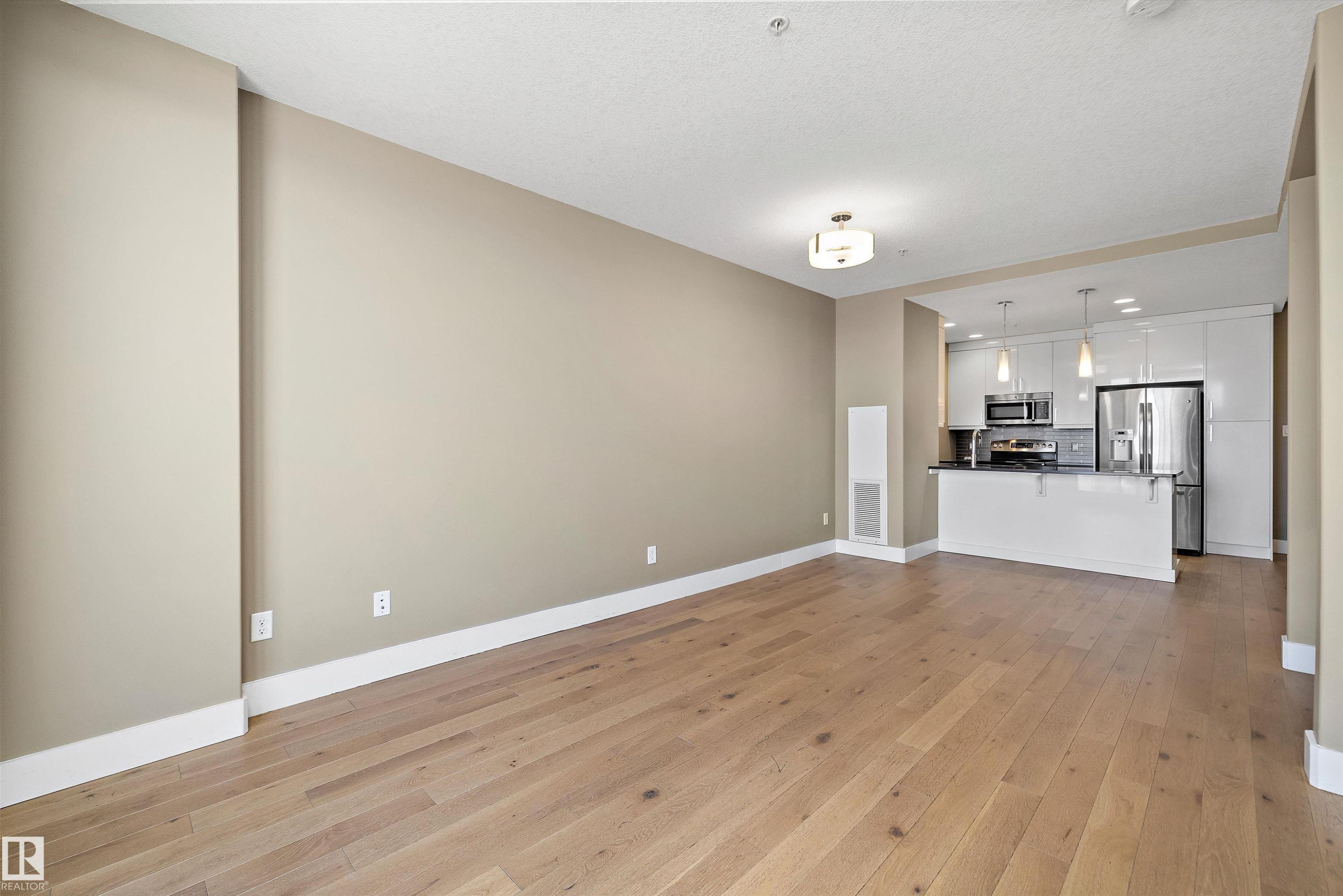 Open-concept living area with wood-finish flooring, a contemporary ceiling light fixture, and neutral wall tones - 307 11080 Ellerslie Rd Sw, Edmonton, AB