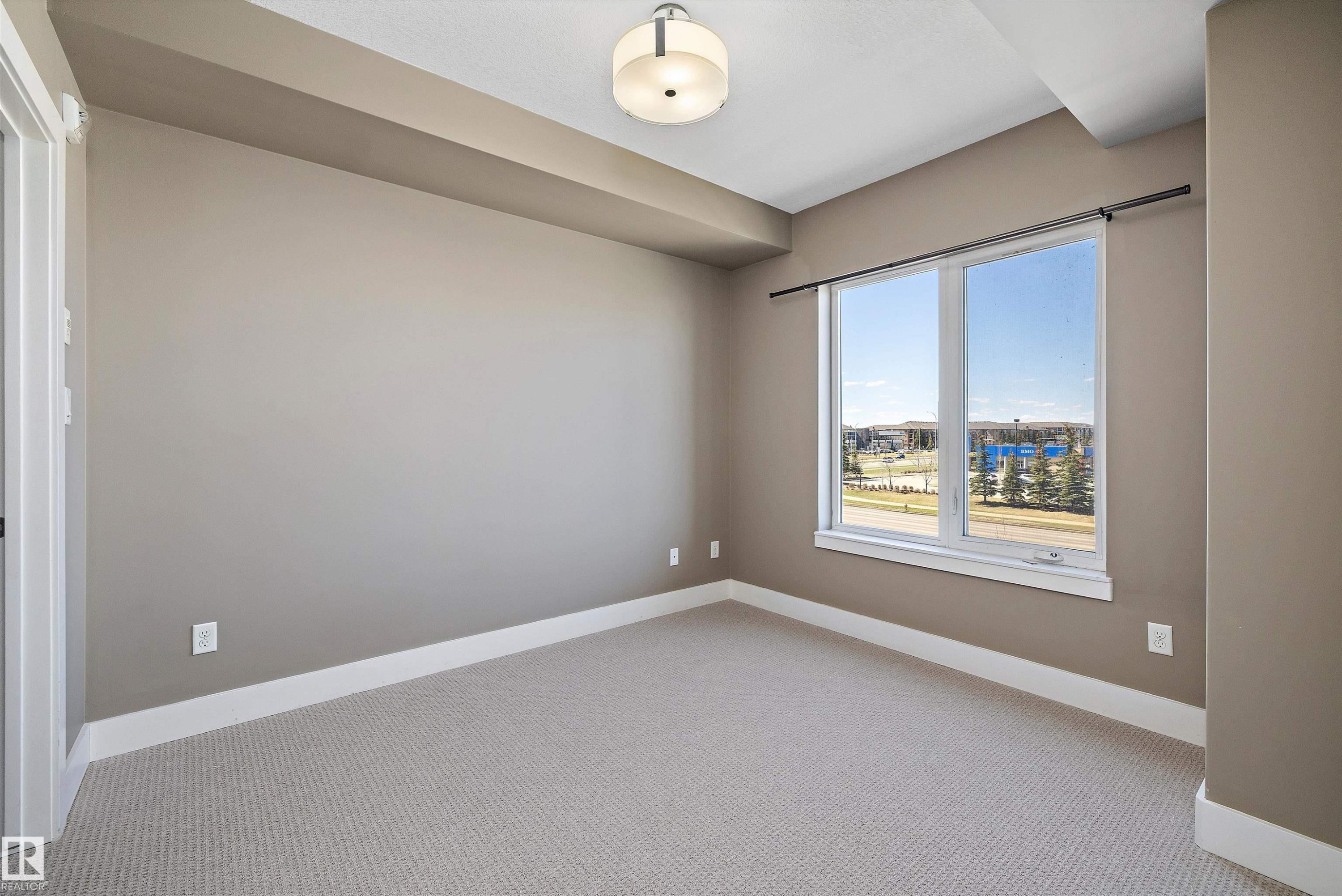 Carpeted room featuring a large window, neutral wall tones, white trim, and a contemporary flush-mount ceiling fixture - 307 11080 Ellerslie Rd Sw, Edmonton, AB