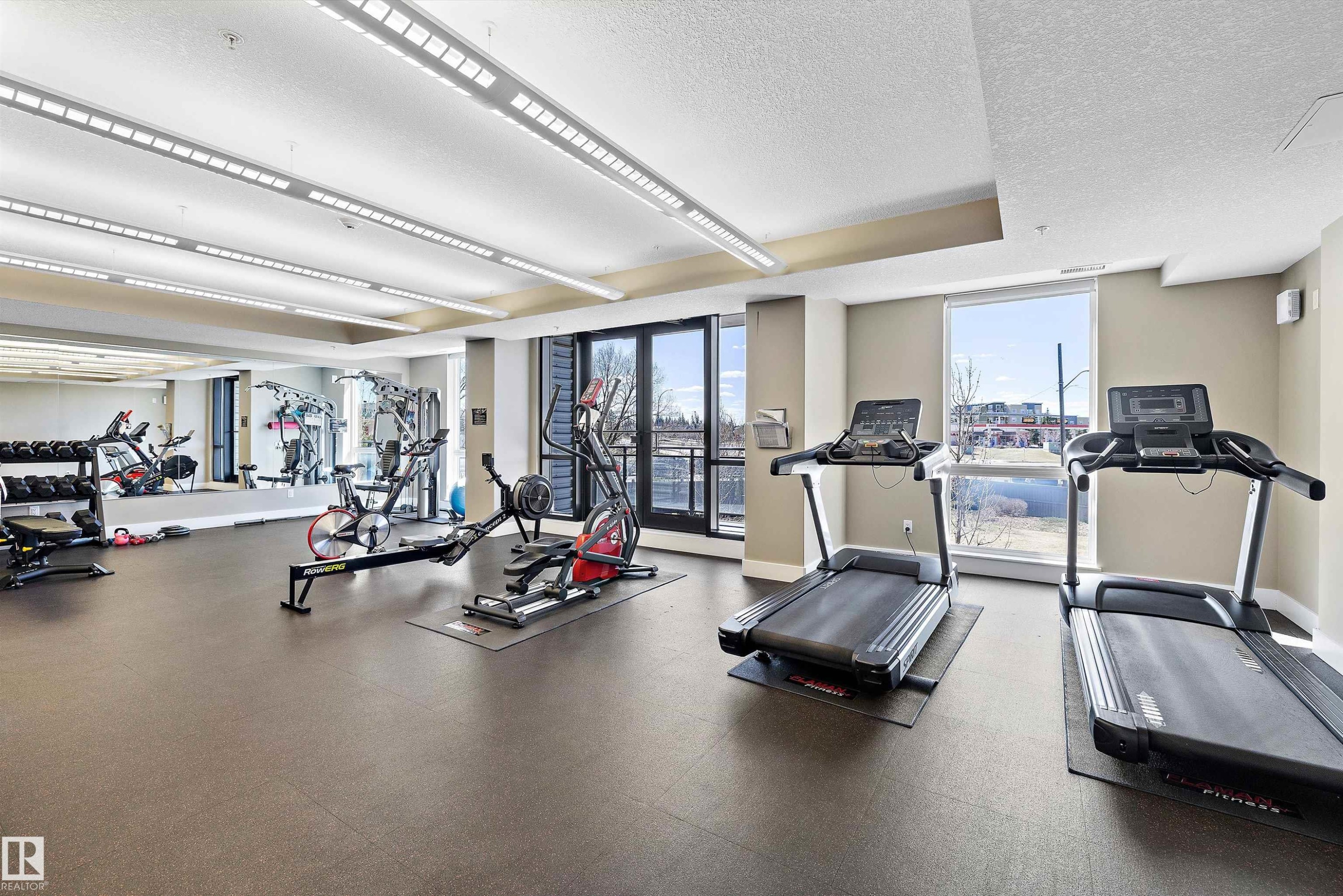 Fitness center featuring rubberized flooring, mirrored wall, multiple cardio machines, and strength training equipment. - 307 11080 Ellerslie Rd Sw, Edmonton, AB