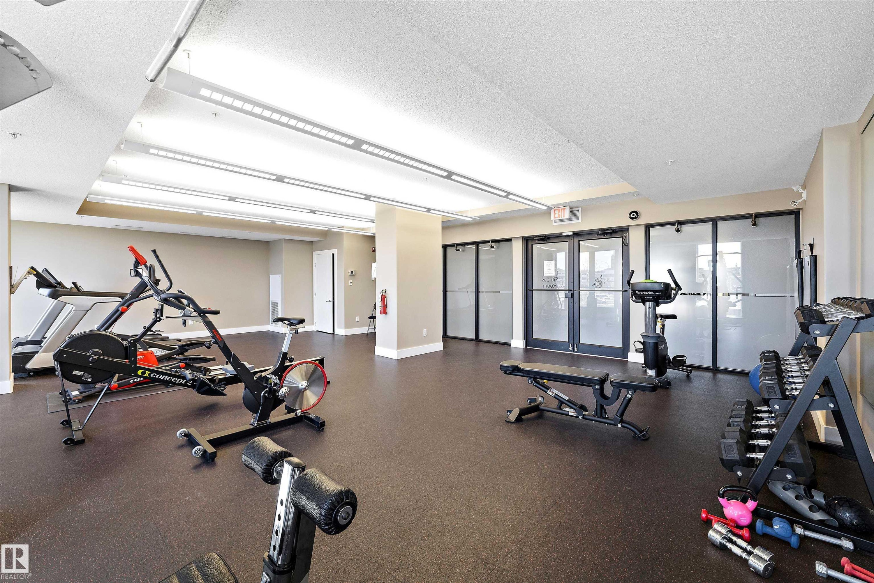 Fitness center featuring rubberized flooring, mirrored wall, multiple cardio machines, and strength training equipment. - 307 11080 Ellerslie Rd Sw, Edmonton, AB