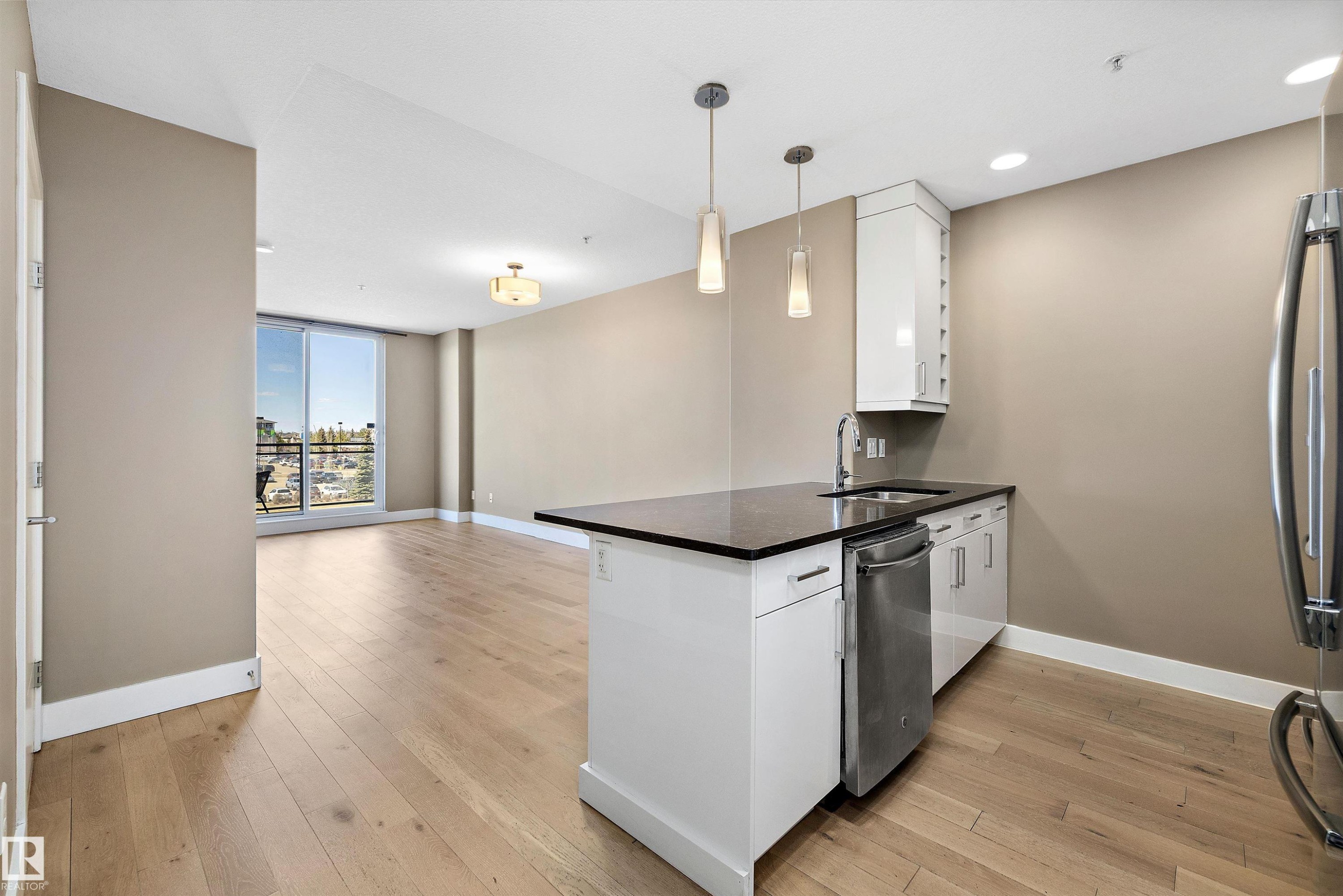 Open-concept living area with wood-finish flooring, a kitchen island featuring a black quartz countertop, and a built-in stainless steel dishwasher. - 307 11080 Ellerslie Rd Sw, Edmonton, AB