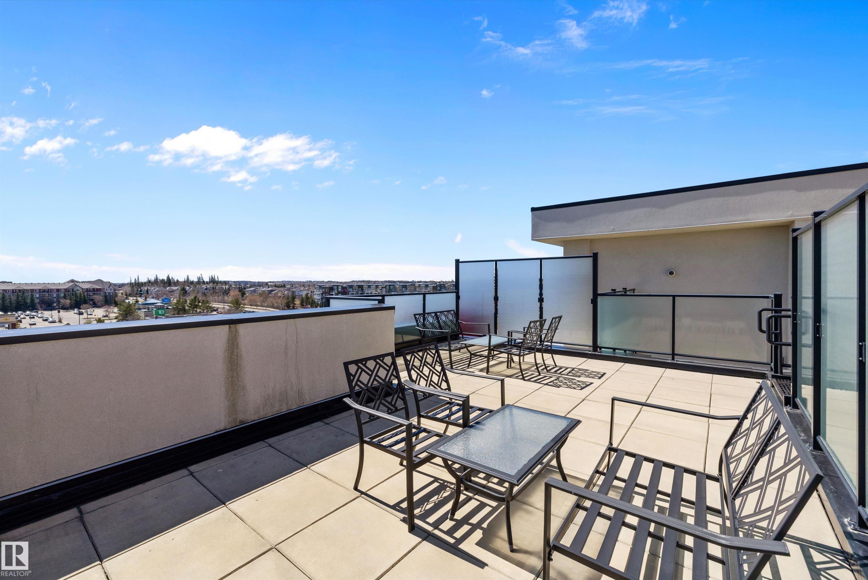 The expansive south rooftop patio featuring large format tile flooring, black metal railing with frosted glass panels, and a surrounding privacy screen, gives an opportunity for a lot of sun and sunsets. - 307 11080 Ellerslie Rd Sw, Edmonton, AB