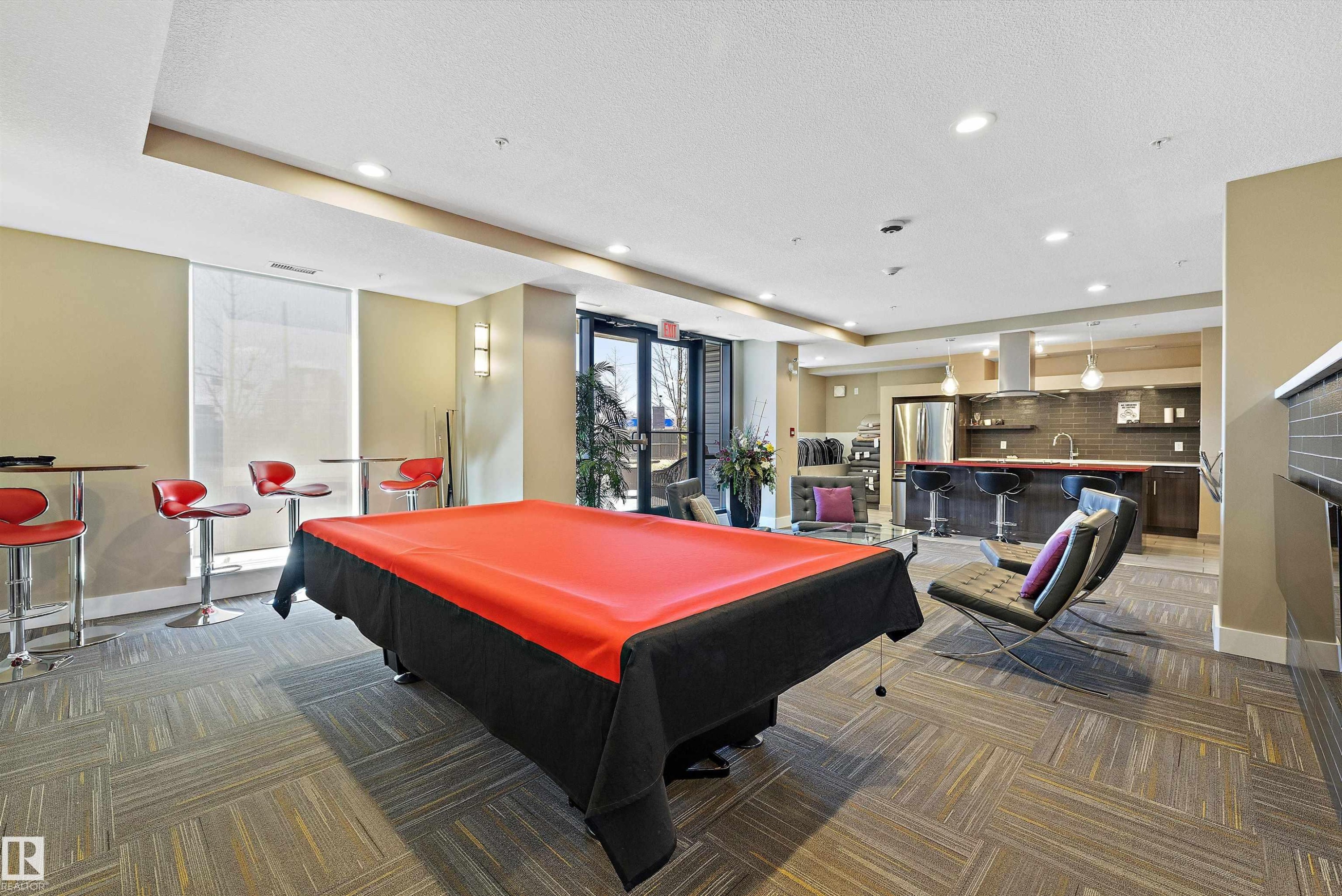 The Recreation Room has a modern kitchen featuring a striking red island countertop, stainless steel appliances, dark wood-finish cabinetry, and a dark subway tile backsplash, with a pool table and a lounge type seating to enjoy with your company. - 307 11080 Ellerslie Rd Sw, Edmonton, AB