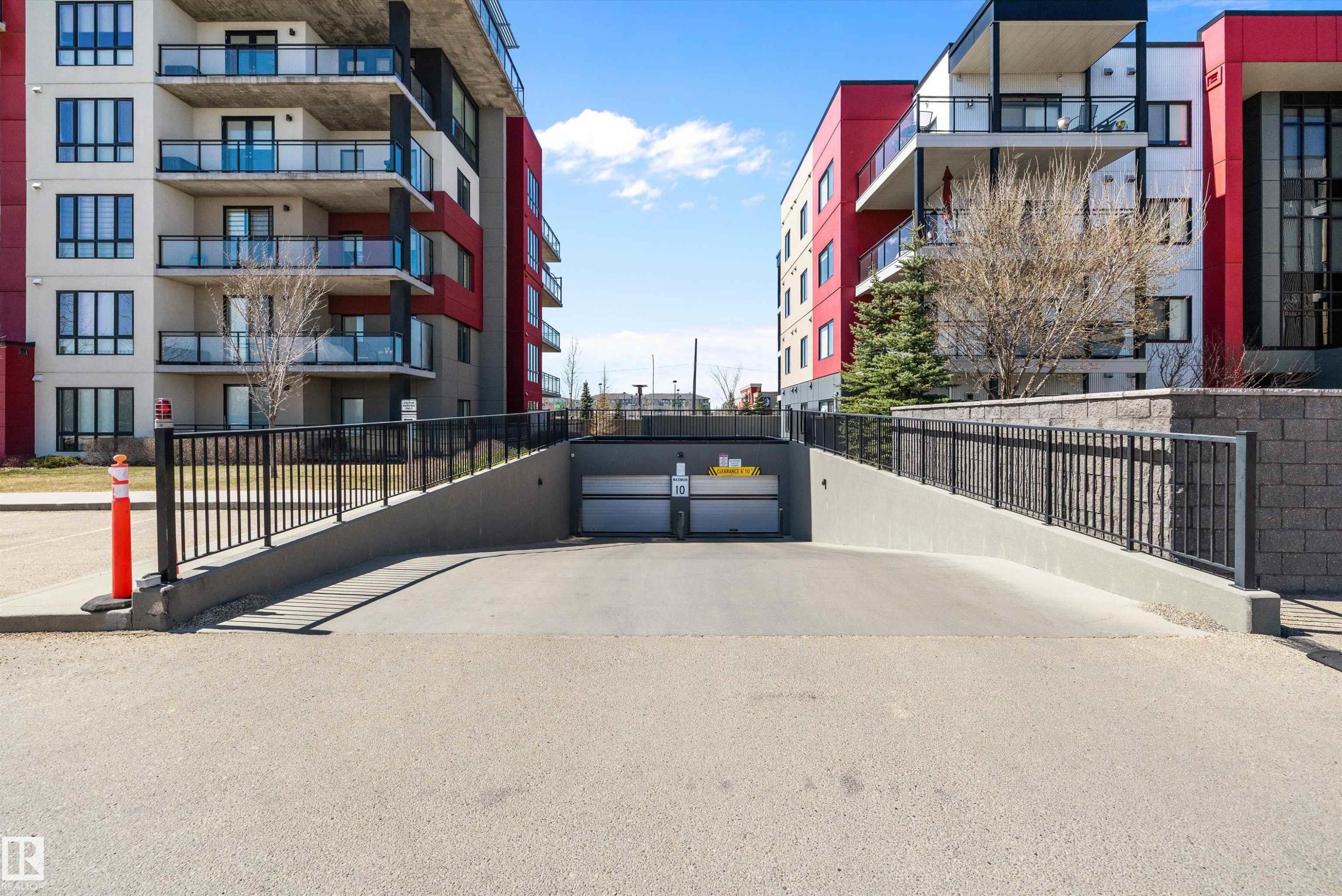 Underground parking access ramp with black metal railings and concrete retaining walls, flanked by two multi-story buildings featuring modern architectural design with red and white facades - 307 11080 Ellerslie Rd Sw, Edmonton, AB