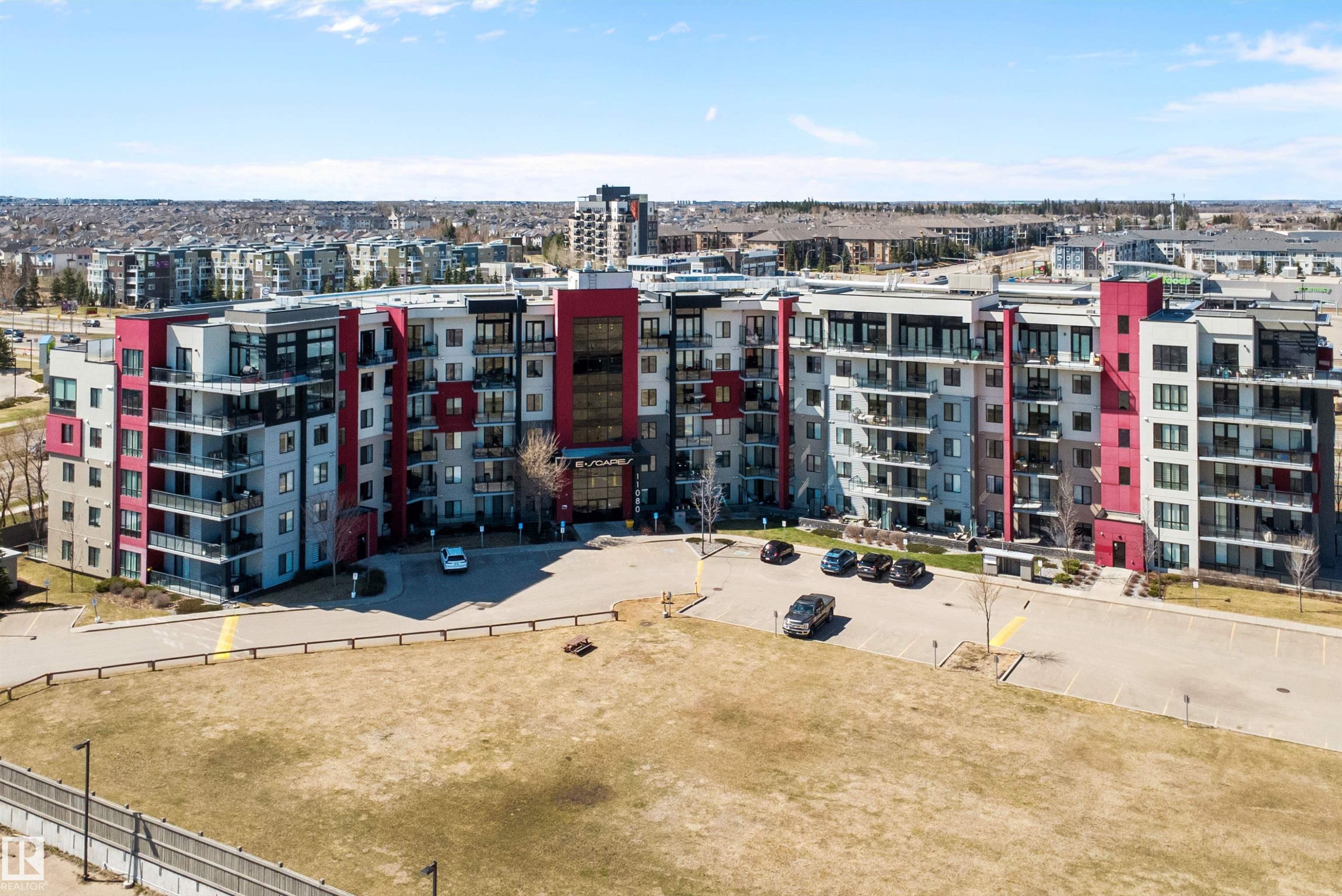 Multi-story residential building featuring a modern facade with red and grey paneling, multiple private balconies, a central entrance, and a paved parking area. - 307 11080 Ellerslie Rd Sw, Edmonton, AB