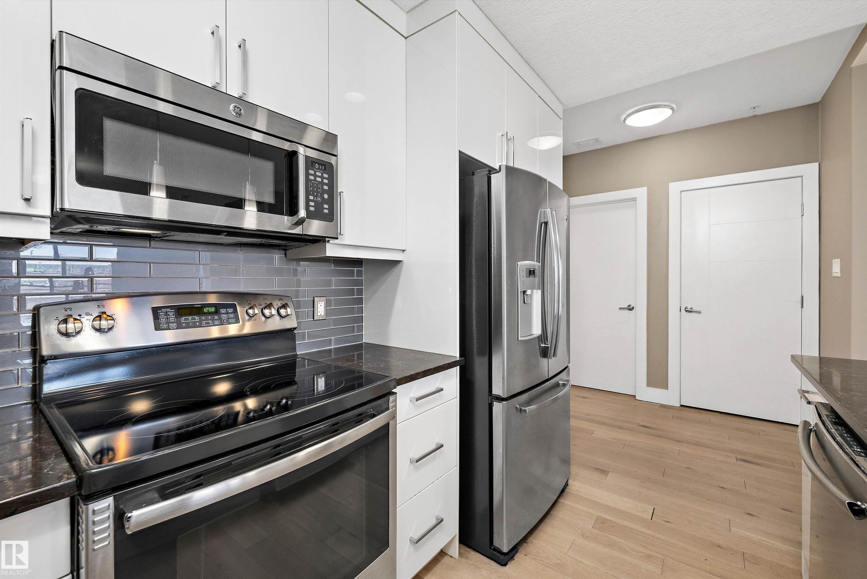Modern kitchen featuring stainless steel appliances, sleek white cabinetry, a subway tile backsplash, dark countertops, and light wood-finish flooring - 307 11080 Ellerslie Rd Sw, Edmonton, AB