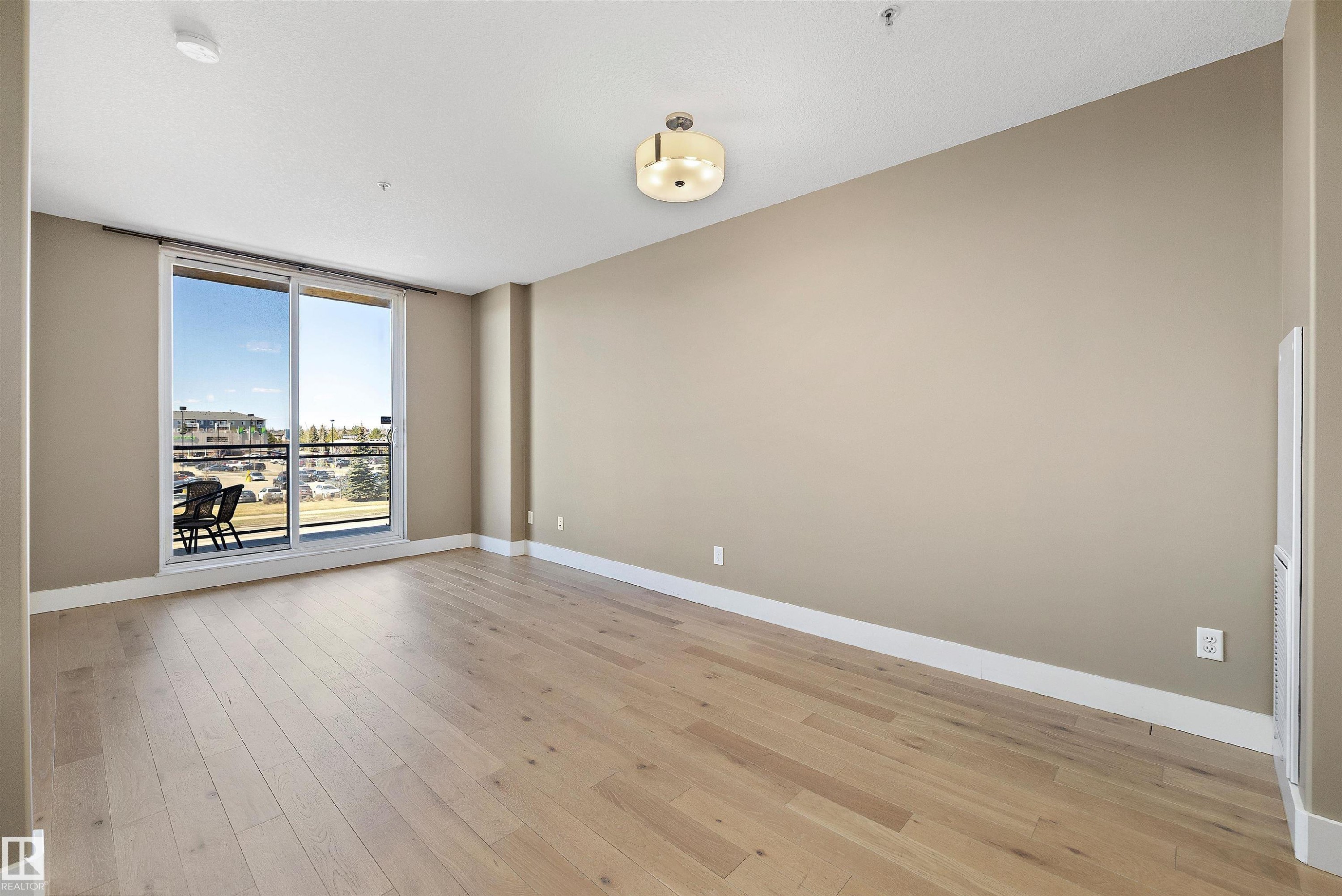 Spacious room featuring wood-finish flooring, a contemporary ceiling light fixture, neutral wall tones, white trim, and a sliding glass door leading to a private balcony with a metal railing. - 307 11080 Ellerslie Rd Sw, Edmonton, AB