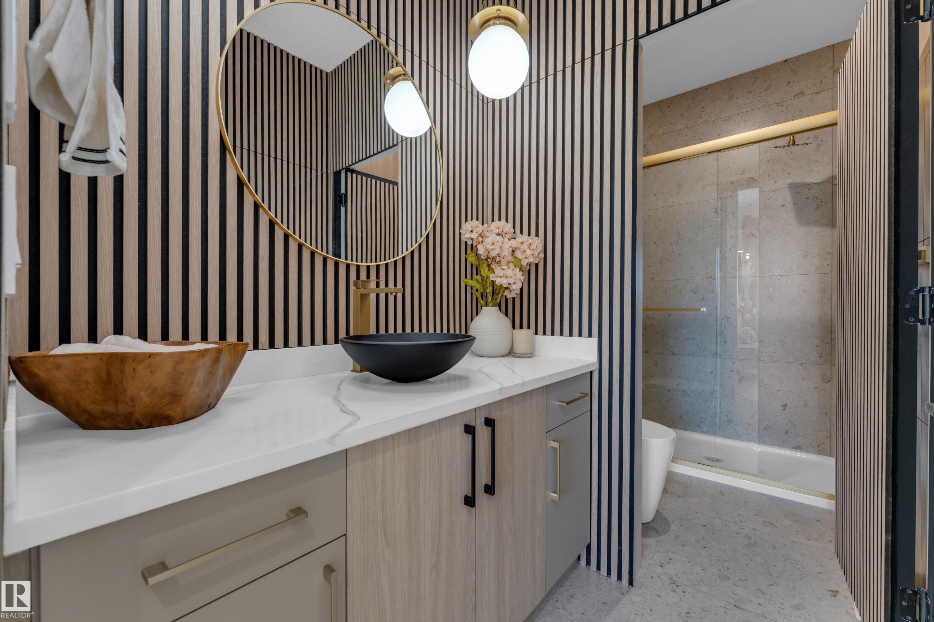 Modern bathroom featuring a vanity with a white countertop and light wood-finish cabinetry, a vessel sink with a gold-finish faucet, and a round gold-framed mirror - 206 Linden Loop, Leduc, AB