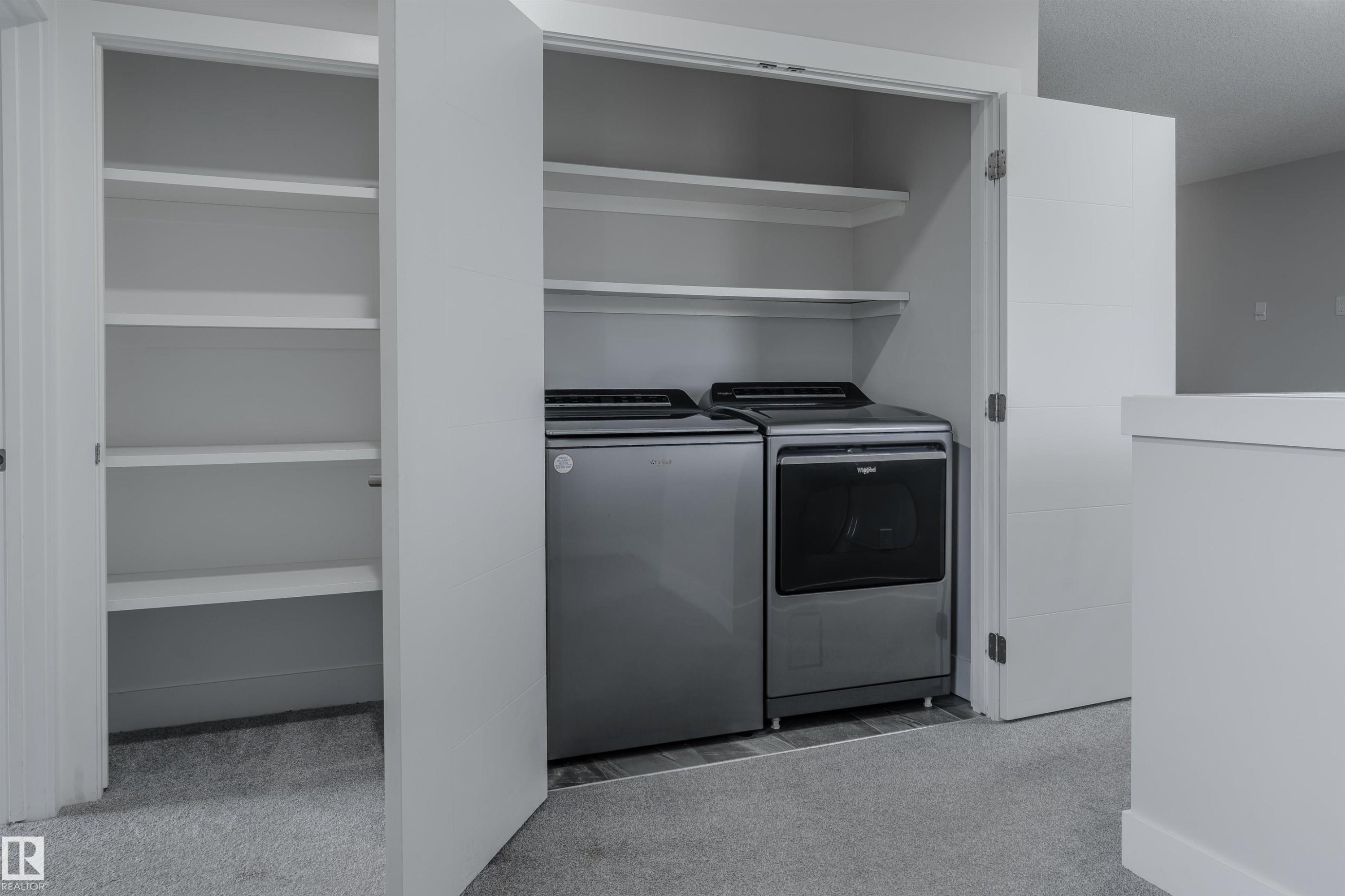Integrated laundry area featuring a side-by-side washer and dryer, built-in shelving, white bifold doors, and light gray carpeting - 2104 160 Street Sw, Edmonton, AB - Indoor Photo Showing Laundry Room