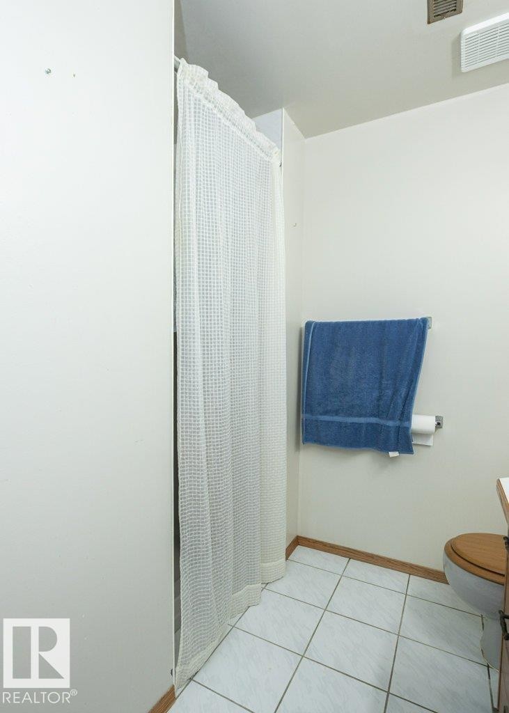 Bathroom featuring a shower with a white waffled curtain, square tile flooring, light wood-finish baseboards, and a toilet with a wood-finish seat - 60230 Rg Rd 271, Rural Westlock County, AB
