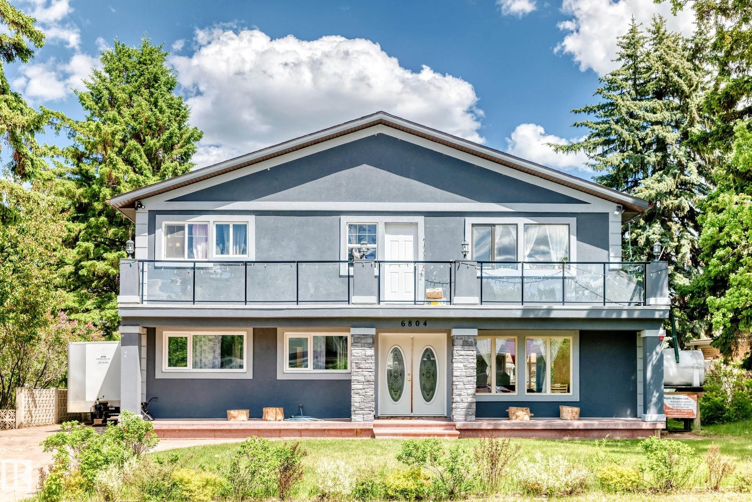 Modern facade featuring a second-story balcony with glass railing, stone accent pillars, double entry doors with oval glass inserts, multiple windows, and a partial front yard - 6804 102A Avenue, Edmonton, AB