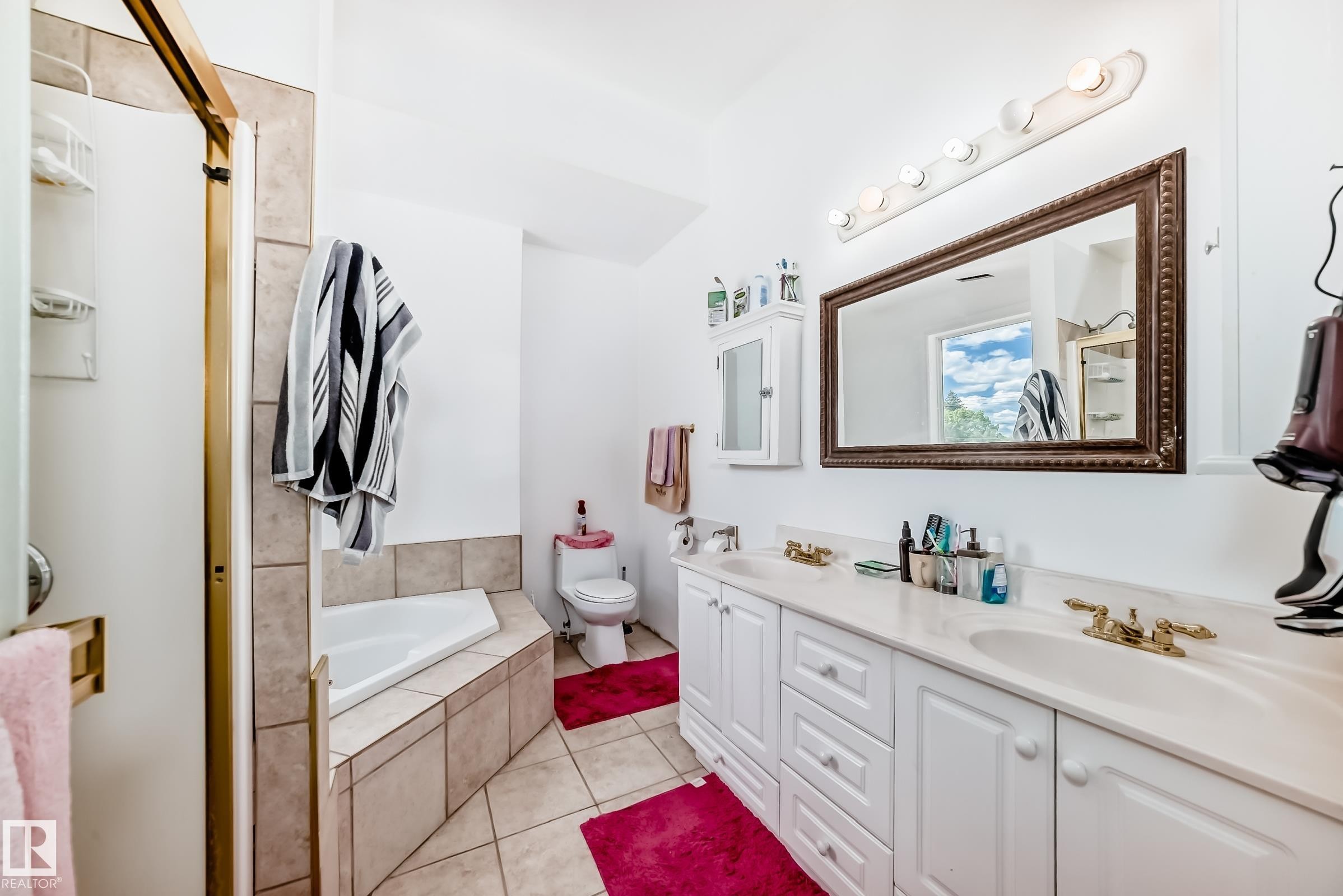 Bathroom featuring a dual vanity with white cabinetry and integrated sinks, a built-in corner bathtub with tiled surround, a separate shower with a gold-toned frame, a wall-mounted medicine cabinet, and a large framed mirror with overhead lighting - 6804 102A Avenue, Edmonton, AB