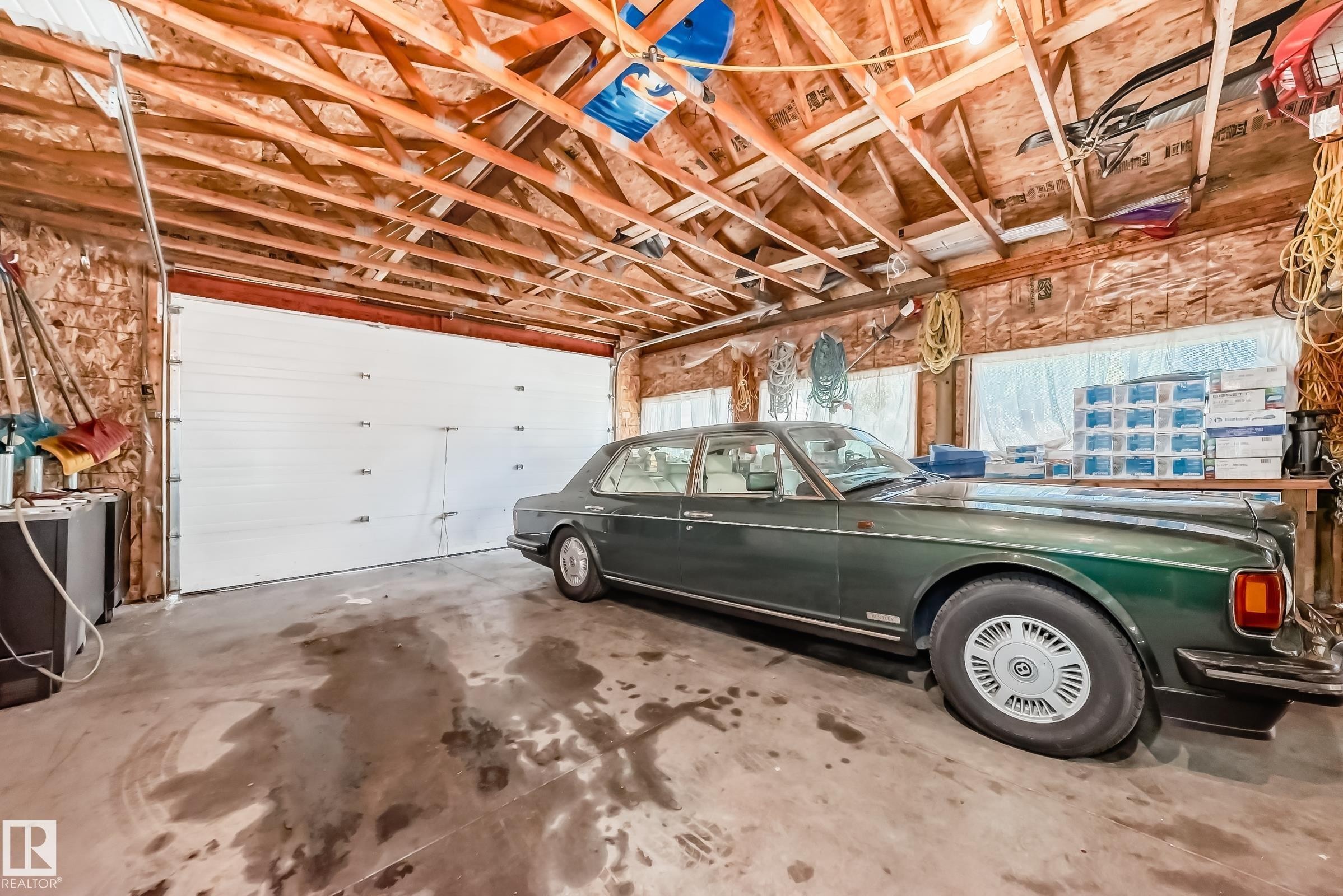 Spacious garage featuring a large white overhead door, exposed wooden ceiling trusses, concrete flooring, windows, and shelving - 6804 102A Avenue, Edmonton, AB