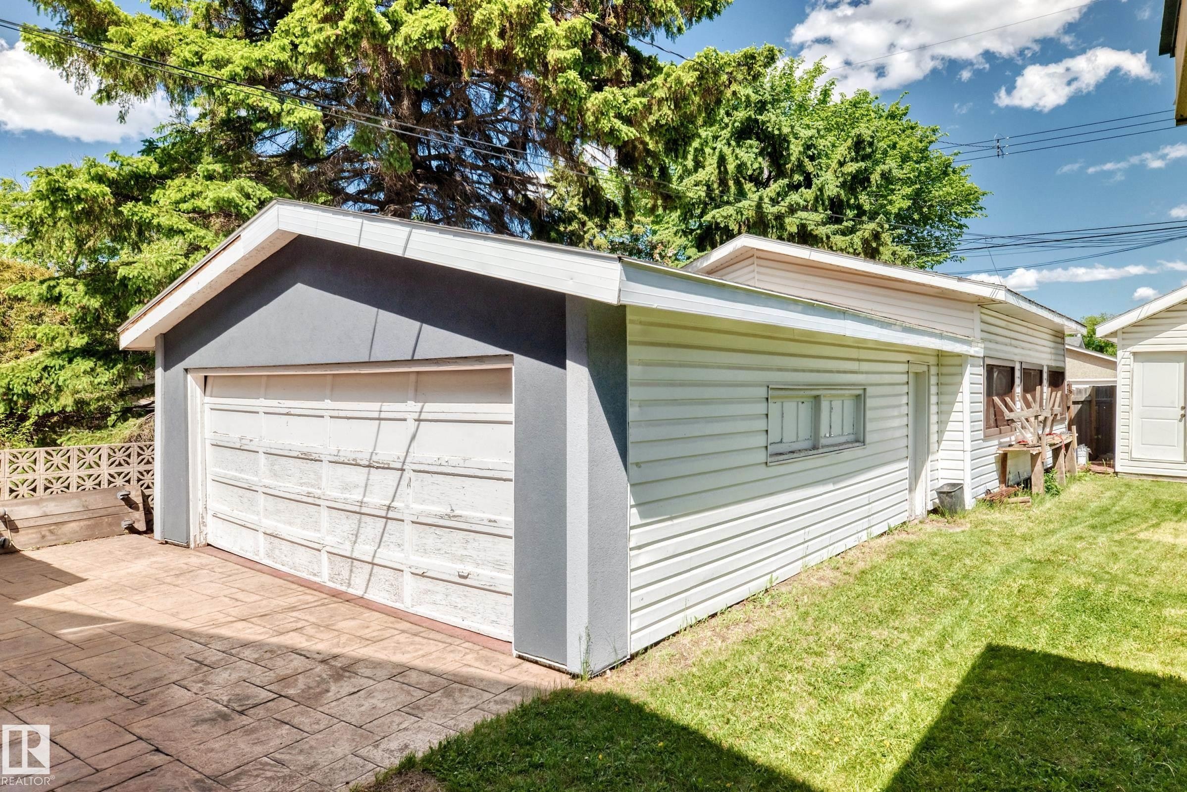 Detached garage featuring a white panel door, light gray stucco facade, white siding, and stamped concrete paving - 6804 102A Avenue, Edmonton, AB