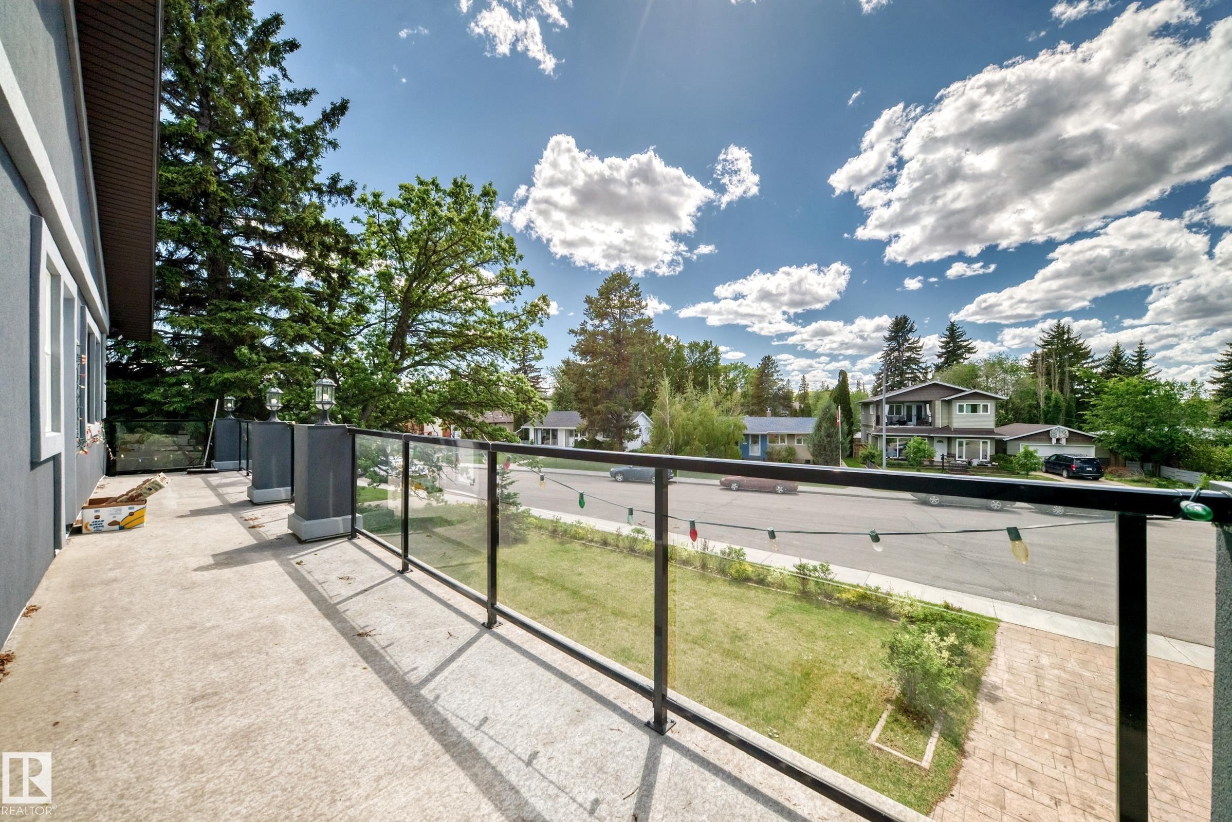 Concrete balcony with a glass and metal railing, featuring a stucco exterior, multiple exterior light fixtures, and a tree-lined streetscape - 6804 102A Avenue, Edmonton, AB