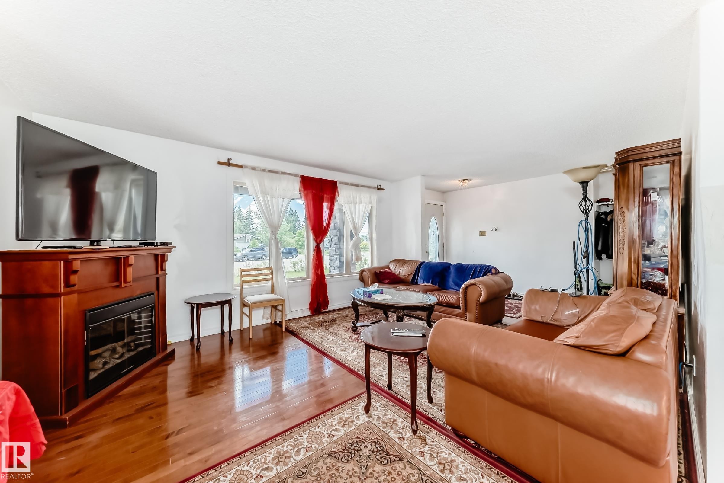 Warm and inviting living area featuring wood-finish flooring, a decorative electric fireplace with a wood-tone mantel, and a large window providing ample natural light - 6804 102A Avenue, Edmonton, AB