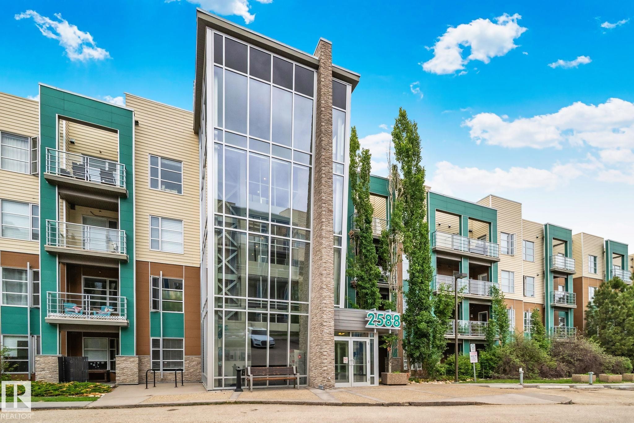 Modern multi-story building featuring a prominent glass-enclosed entrance, stacked stone accents, and varied siding in shades of beige and teal - 217 2588 Anderson Way, Edmonton, AB