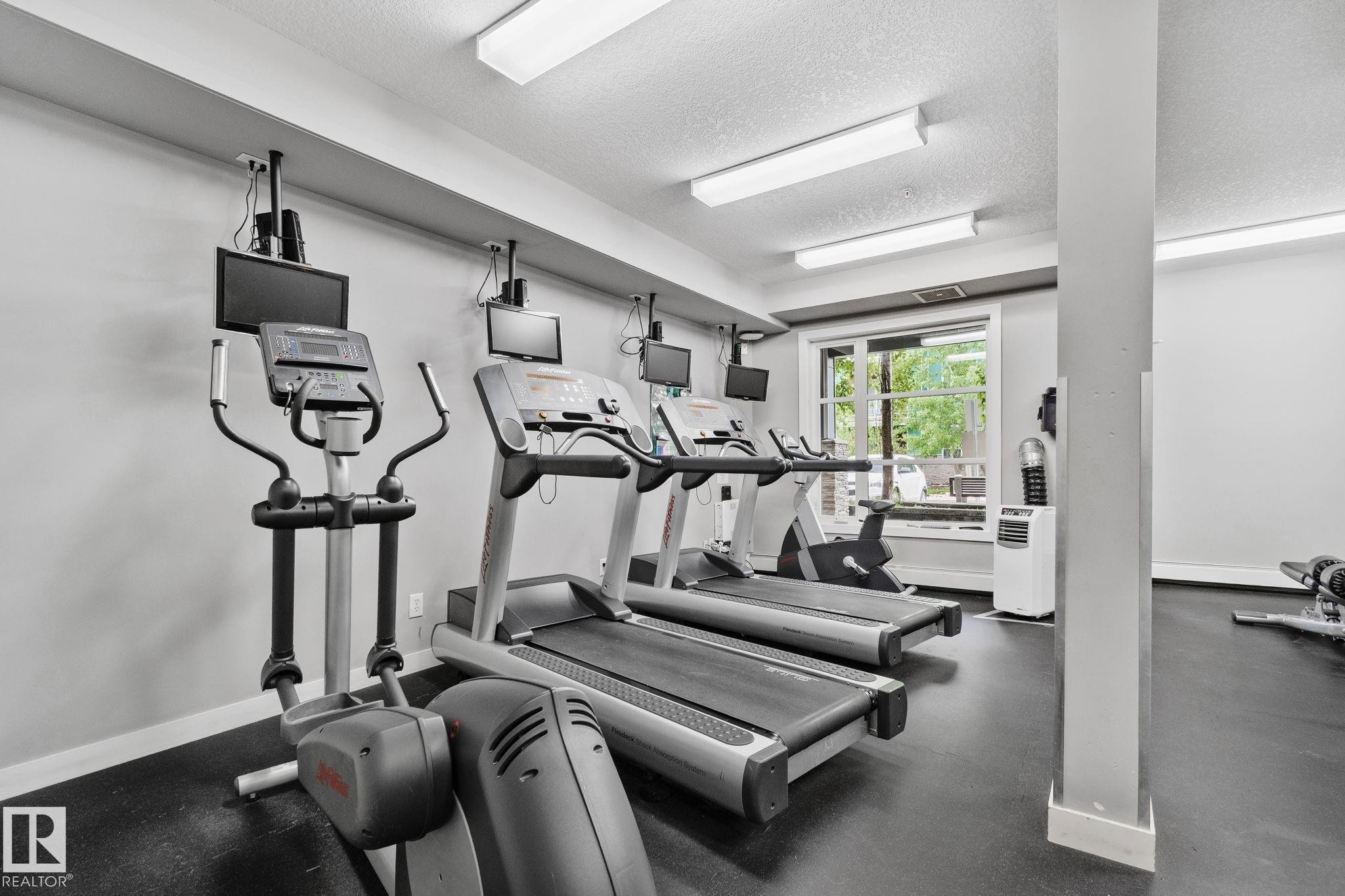 Fitness room featuring multiple cardio machines, ceiling-mounted monitors, overhead fluorescent lighting, a window with exterior views, and durable black rubber flooring - 217 2588 Anderson Way, Edmonton, AB