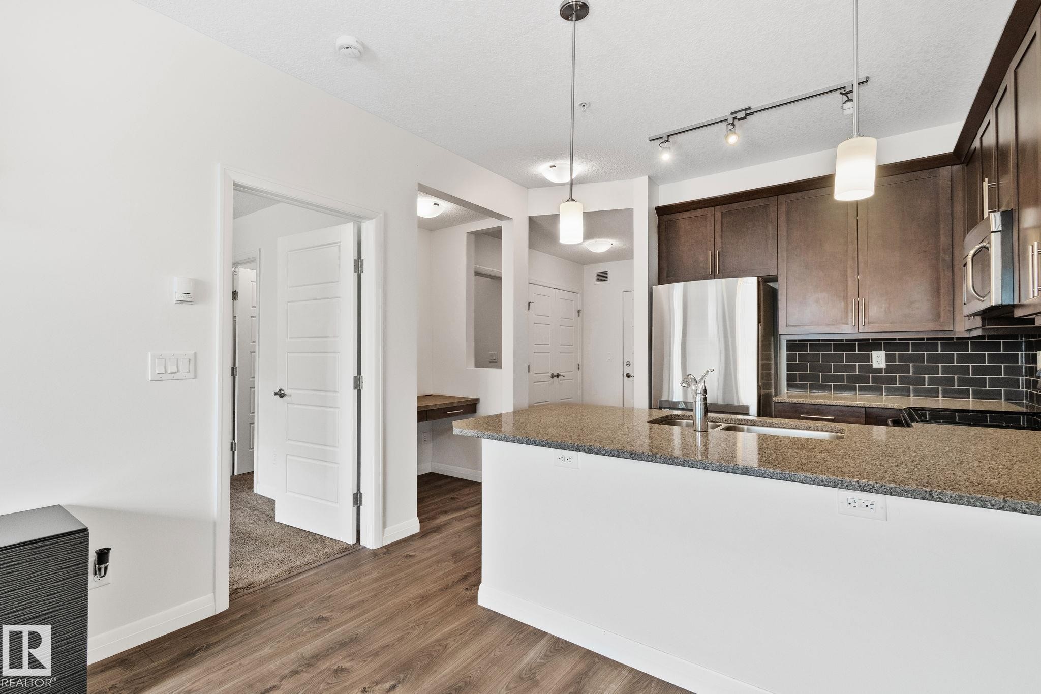 Kitchen island featuring a light-toned stone countertop, integrated sink, and modern pendant lighting - 217 2588 Anderson Way, Edmonton, AB