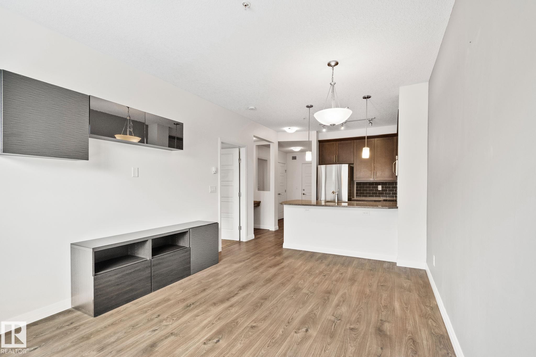 Open-concept living area with wood-finish flooring, featuring a built-in media console, an integrated kitchen with stainless steel appliances, dark wood cabinetry, and a subway tile backsplash - 217 2588 Anderson Way, Edmonton, AB