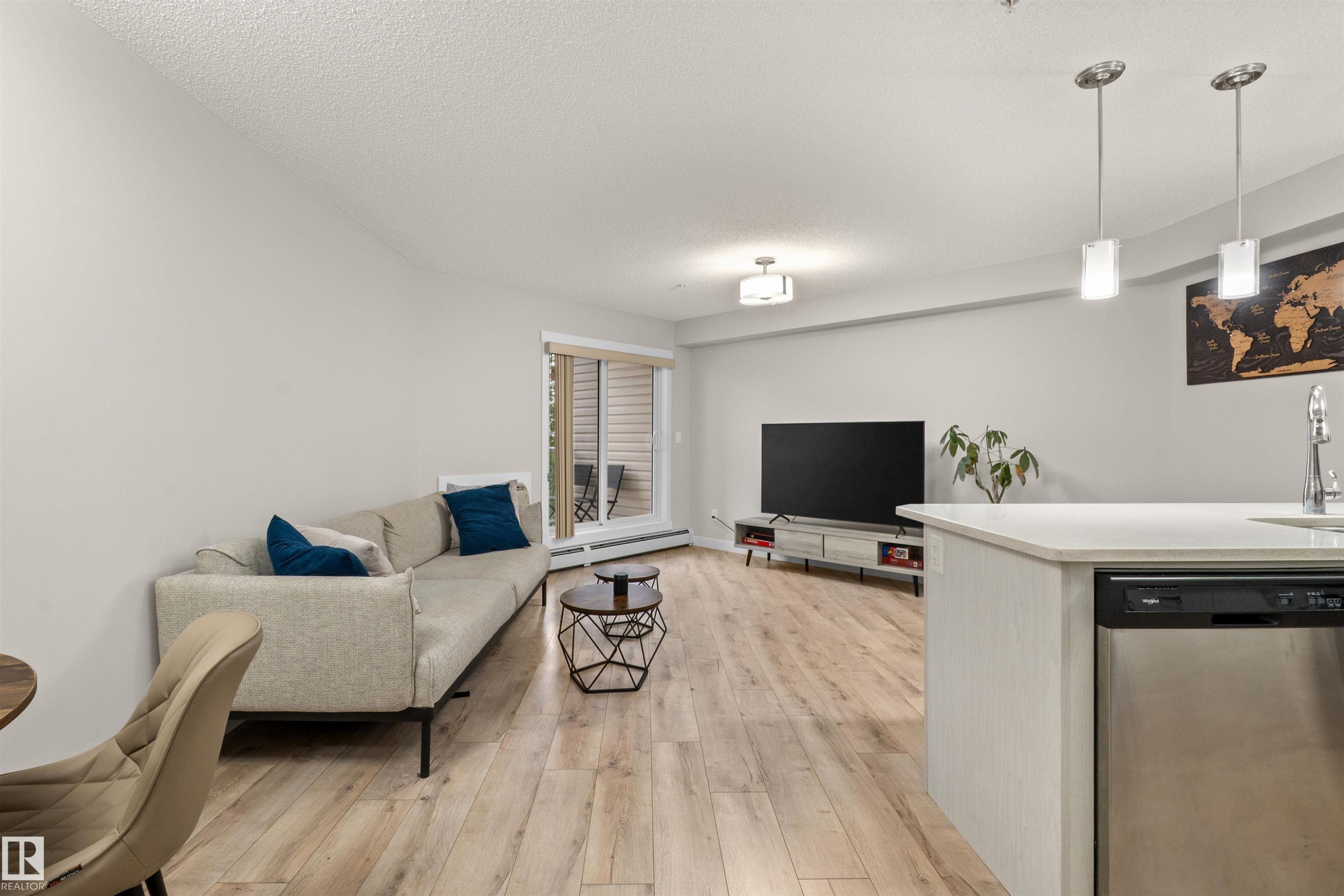 Open-concept living area with wood-finish flooring, a contemporary ceiling light fixture, and a sliding glass door leading to an outdoor space - 210 2229 44 Avenue, Edmonton, AB
