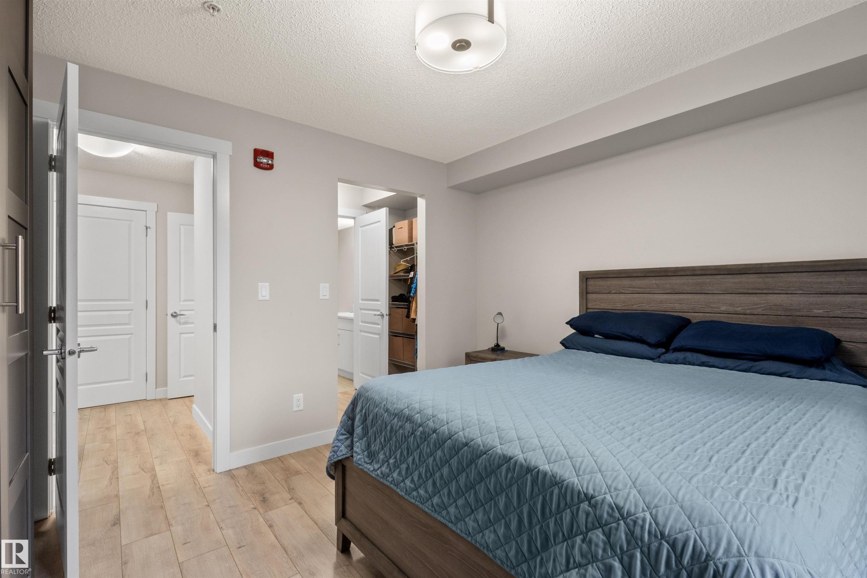 Light wood-finish flooring extends through this room, featuring a wood-panel headboard, a white ceiling with a flush-mount light fixture, and white trim - 210 2229 44 Avenue, Edmonton, AB