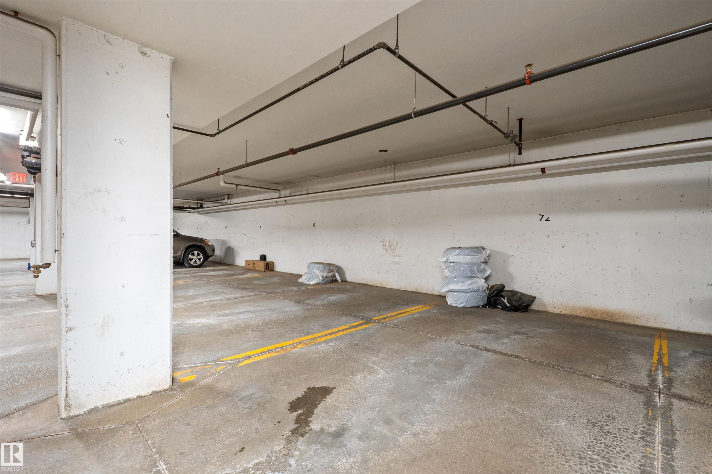 Concrete parking area featuring painted yellow lines, exposed overhead piping, and a white concrete support column - 210 2229 44 Avenue, Edmonton, AB