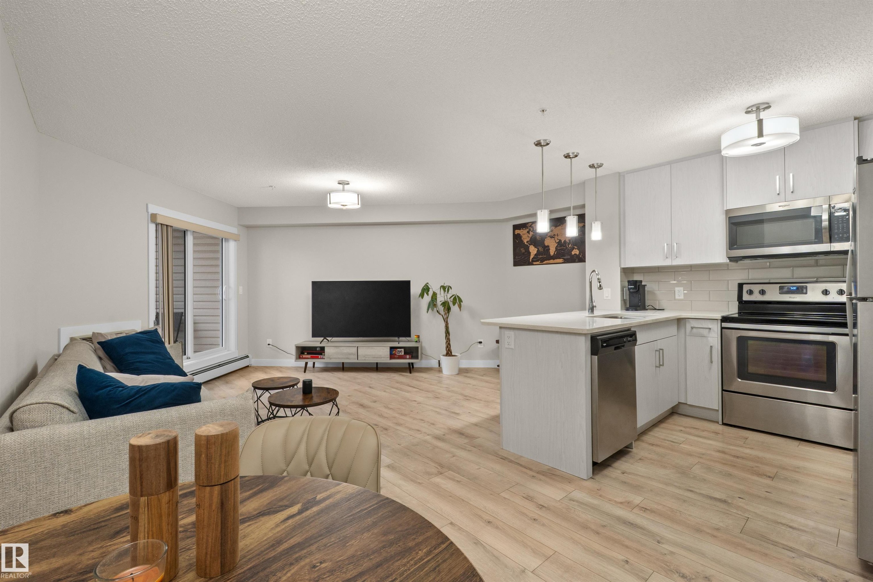 Open-concept living space featuring wood-finish flooring, a kitchen island with pendant lighting, stainless steel appliances, white cabinetry, and a sliding glass door to an outdoor area - 210 2229 44 Avenue, Edmonton, AB