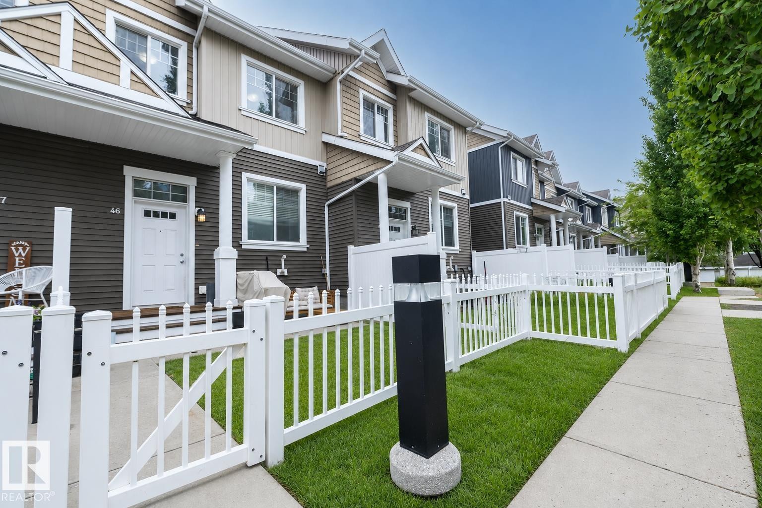 Townhome exterior featuring a white picket fence, a small front yard with green grass, a concrete sidewalk, and a white entry door with frosted glass inserts - 46 4050 Savaryn Drive, Edmonton, AB