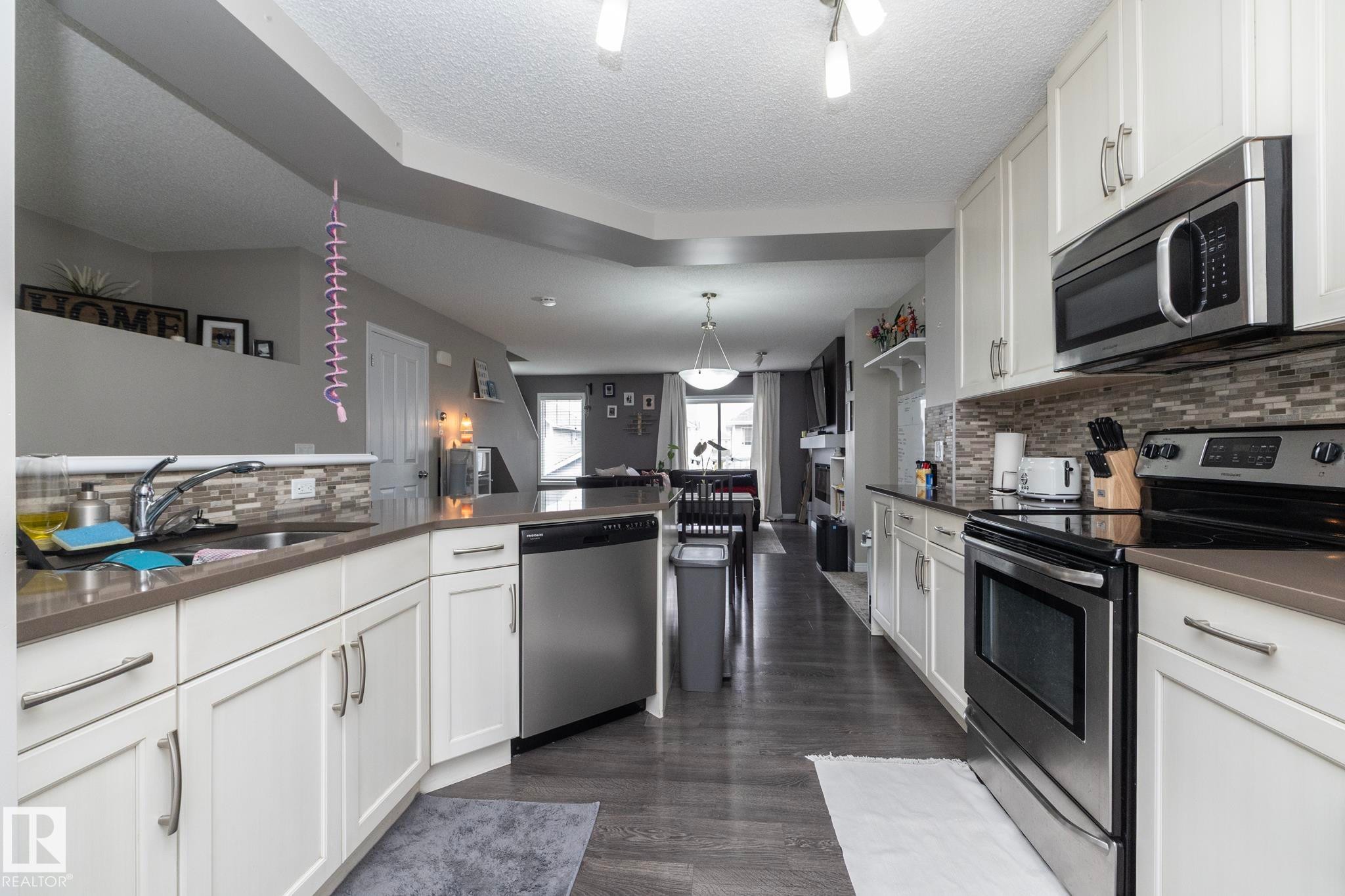Kitchen featuring white shaker-style cabinetry, stainless steel appliances, dark wood-finish flooring, and light-toned countertops with a mosaic tile backsplash - 46 4050 Savaryn Drive, Edmonton, AB