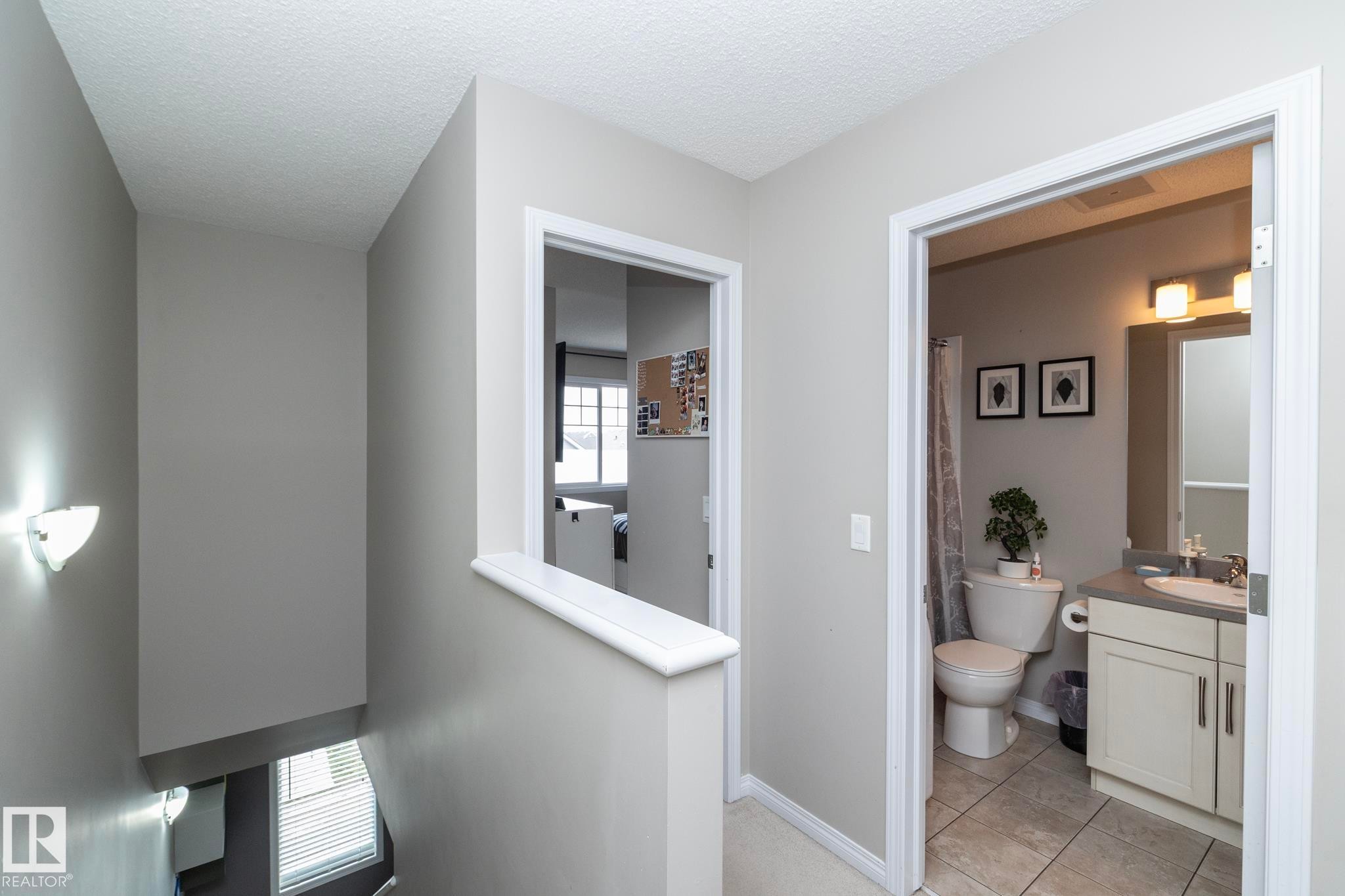 Hallway featuring a wall-mounted sconce, leading to a bathroom with a single vanity, a toilet, and tiled flooring - 46 4050 Savaryn Drive, Edmonton, AB
