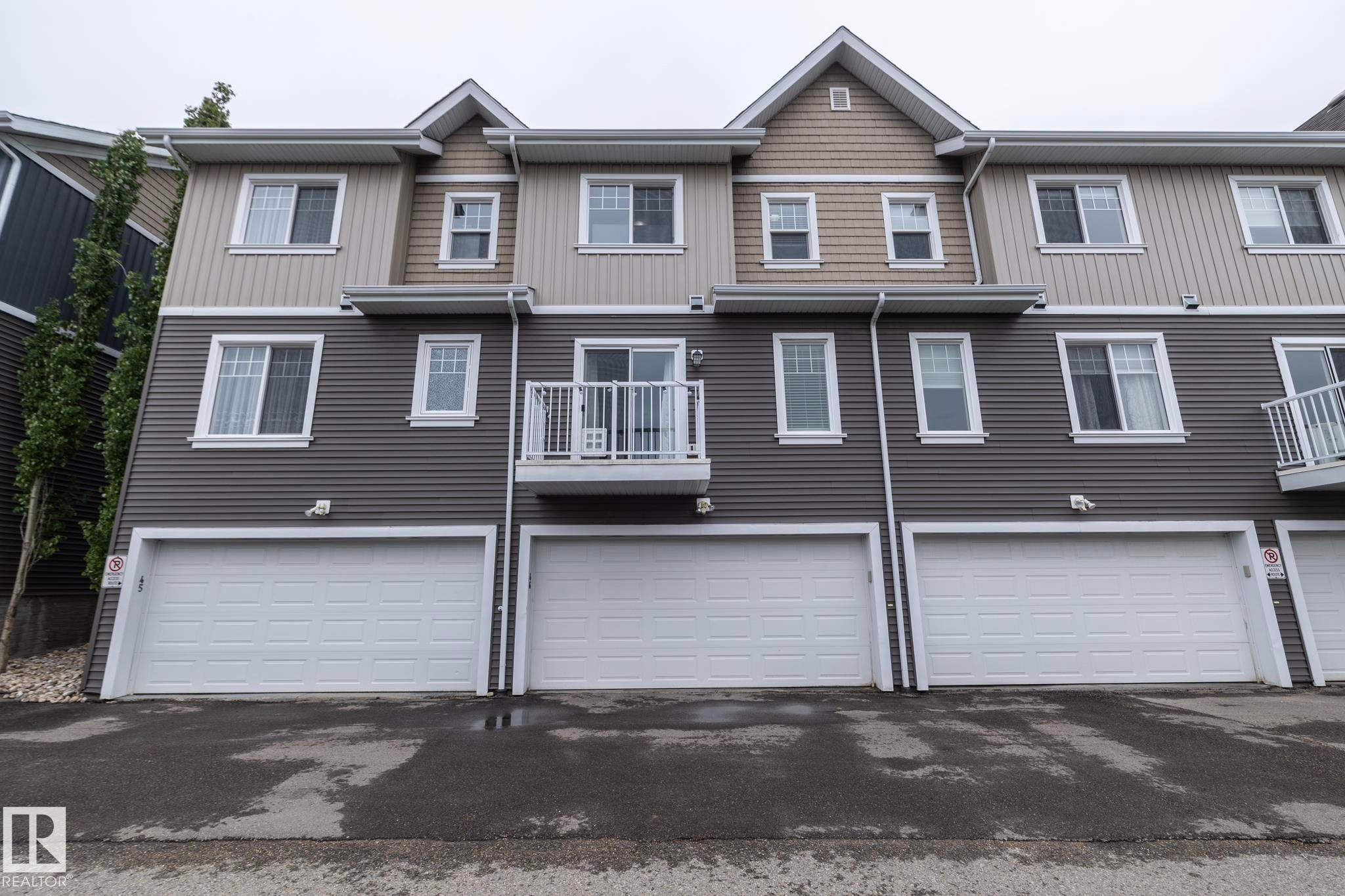 Multi-level exterior featuring grey horizontal siding, tan shake accents, white-trimmed windows, and three individual garages with white panel doors - 46 4050 Savaryn Drive, Edmonton, AB