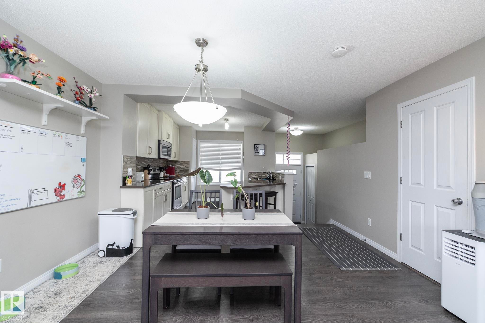 Open-concept living area featuring dark wood-finish flooring, a contemporary dining fixture, and kitchen with light cabinetry, dark countertops, and stainless steel appliances - 46 4050 Savaryn Drive, Edmonton, AB