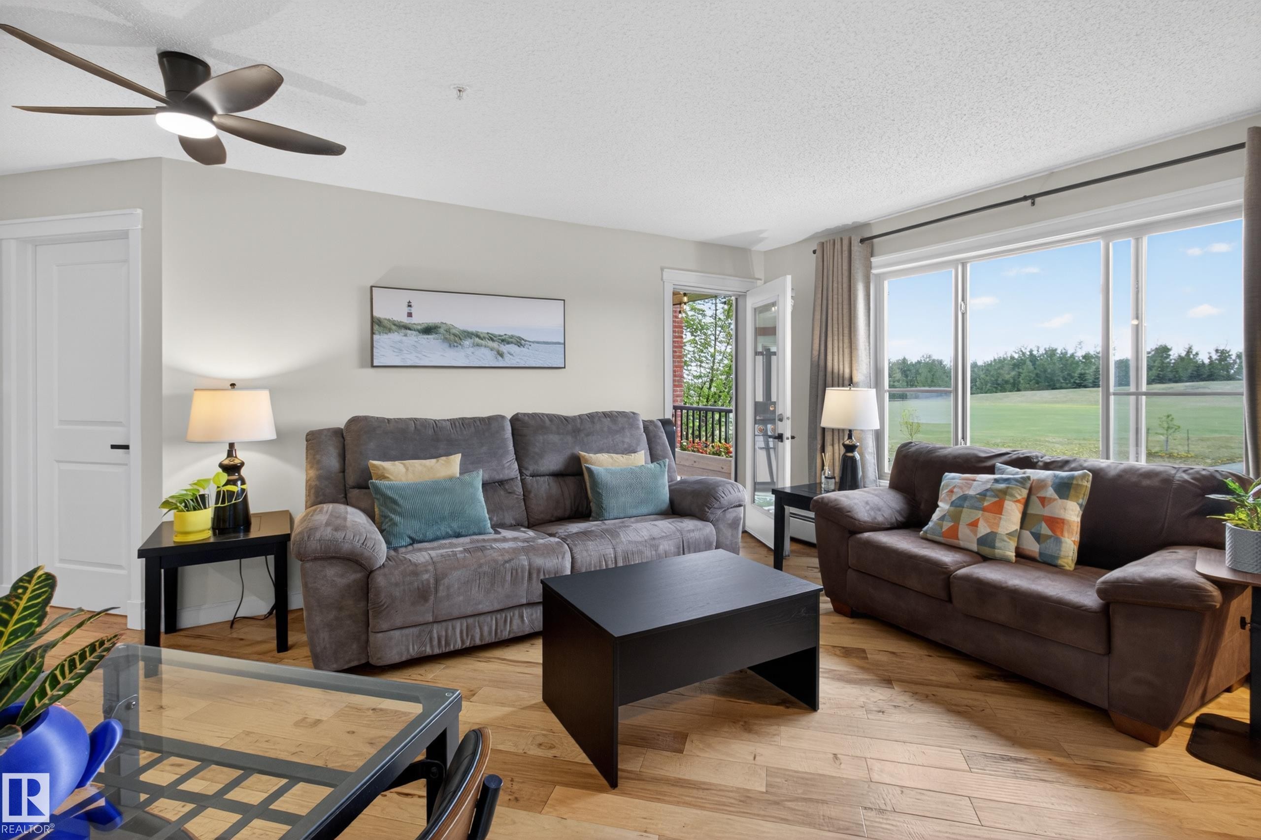 Spacious living area featuring large windows, wood-finish flooring, a ceiling fan, and access to an outdoor balcony - 211 3719 Whitelaw Lane, Edmonton, AB