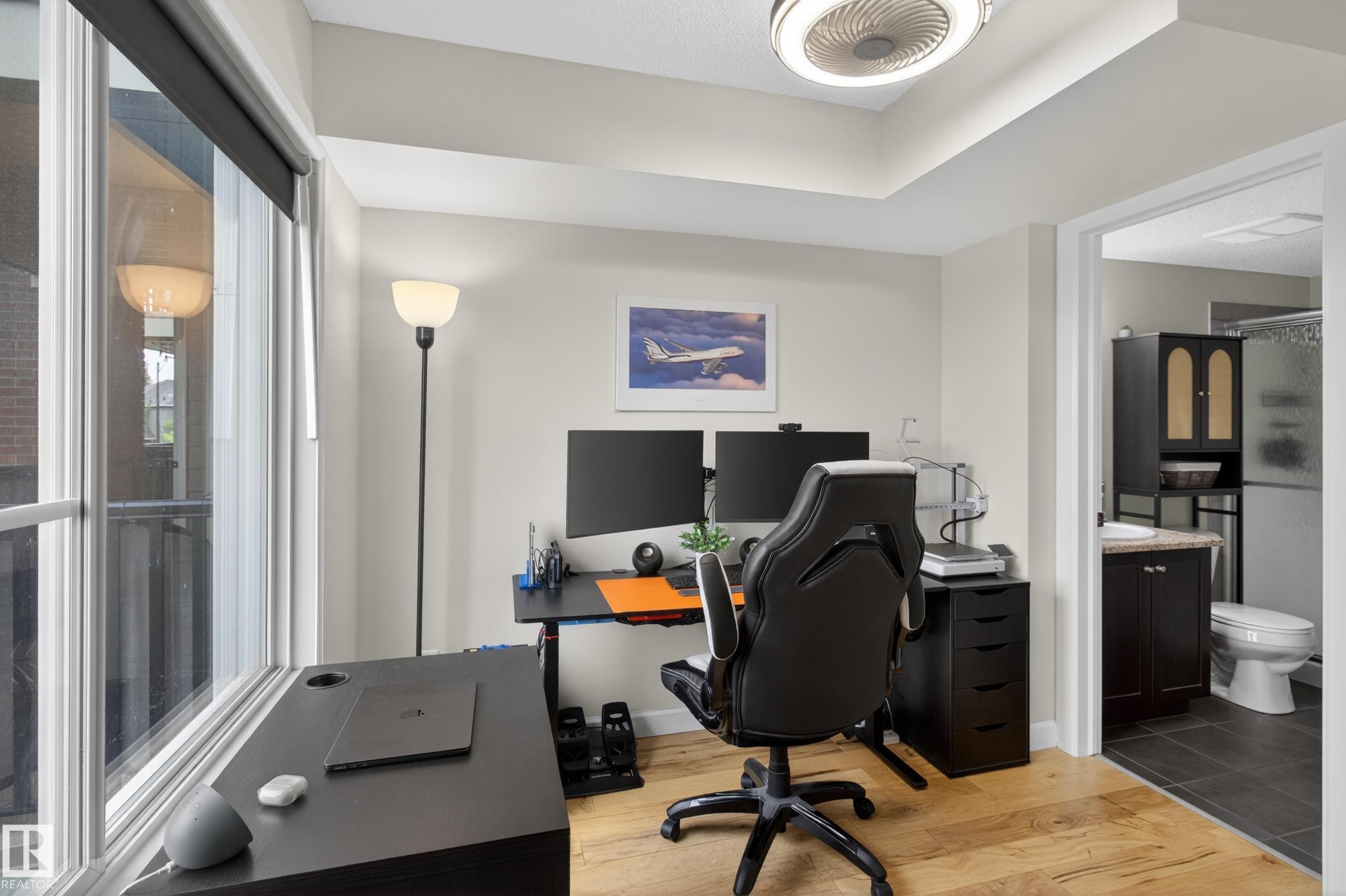 Spacious room featuring light wood-finish flooring, a tray ceiling with a modern flush-mount light fixture, and an expansive window with a roller shade - 211 3719 Whitelaw Lane, Edmonton, AB