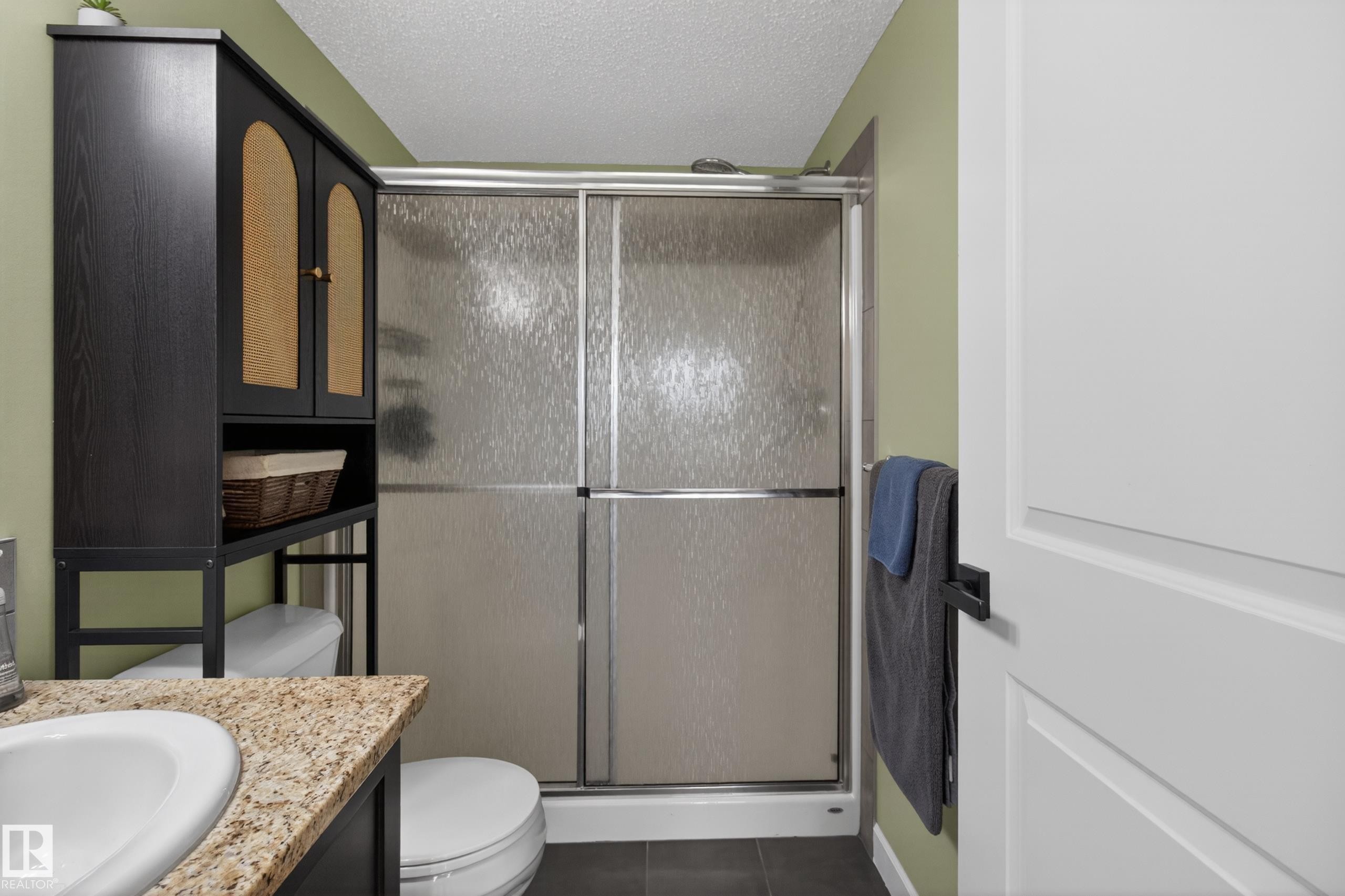 Bathroom featuring a shower with textured glass enclosure, a vanity with a light-colored speckled countertop, an integrated sink, and an over-the-toilet storage cabinet with textured door inserts - 211 3719 Whitelaw Lane, Edmonton, AB