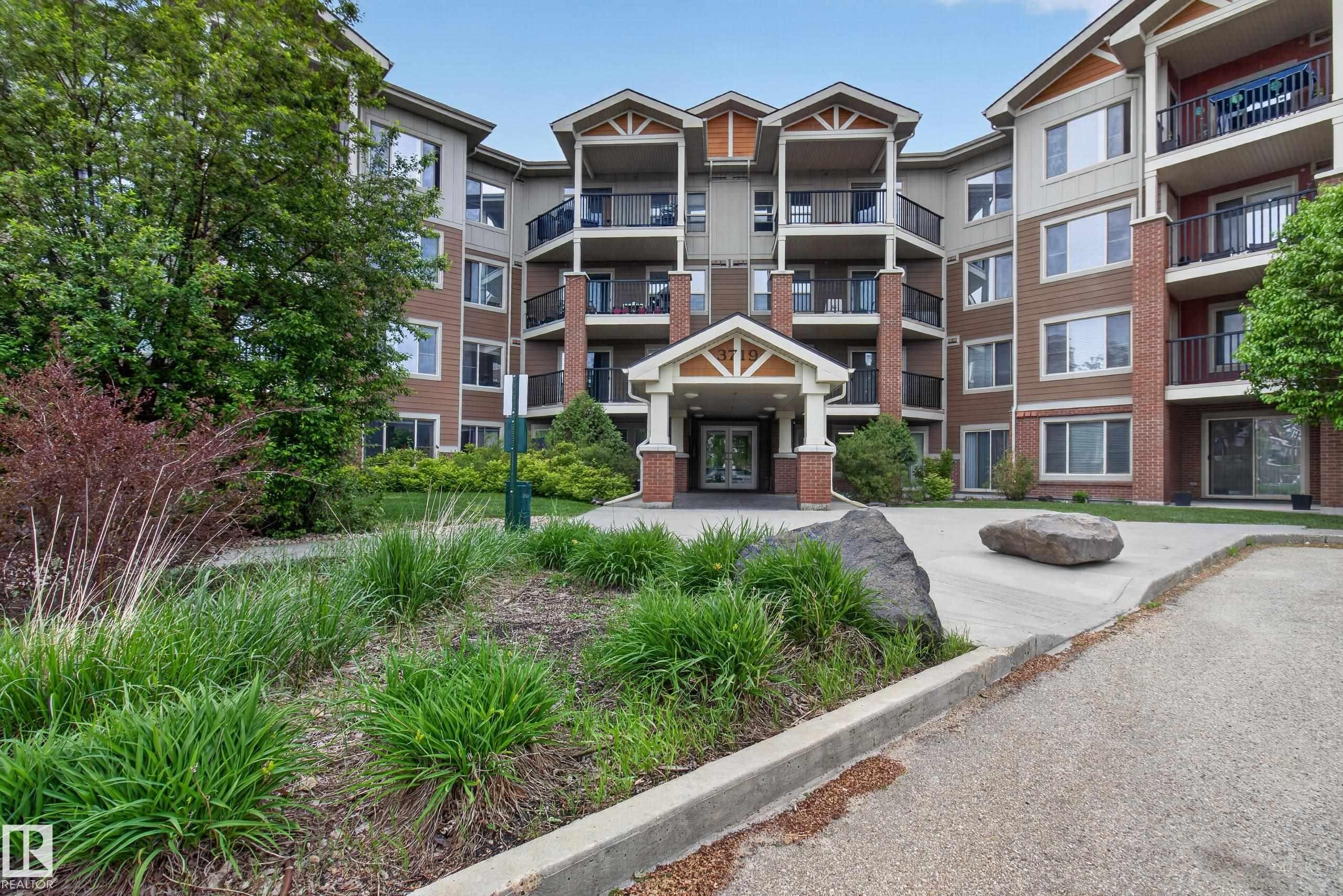 Multi-story building featuring brick and siding facade, multiple balconies with dark railings, a covered entrance, and surrounding landscaping - 211 3719 Whitelaw Lane, Edmonton, AB
