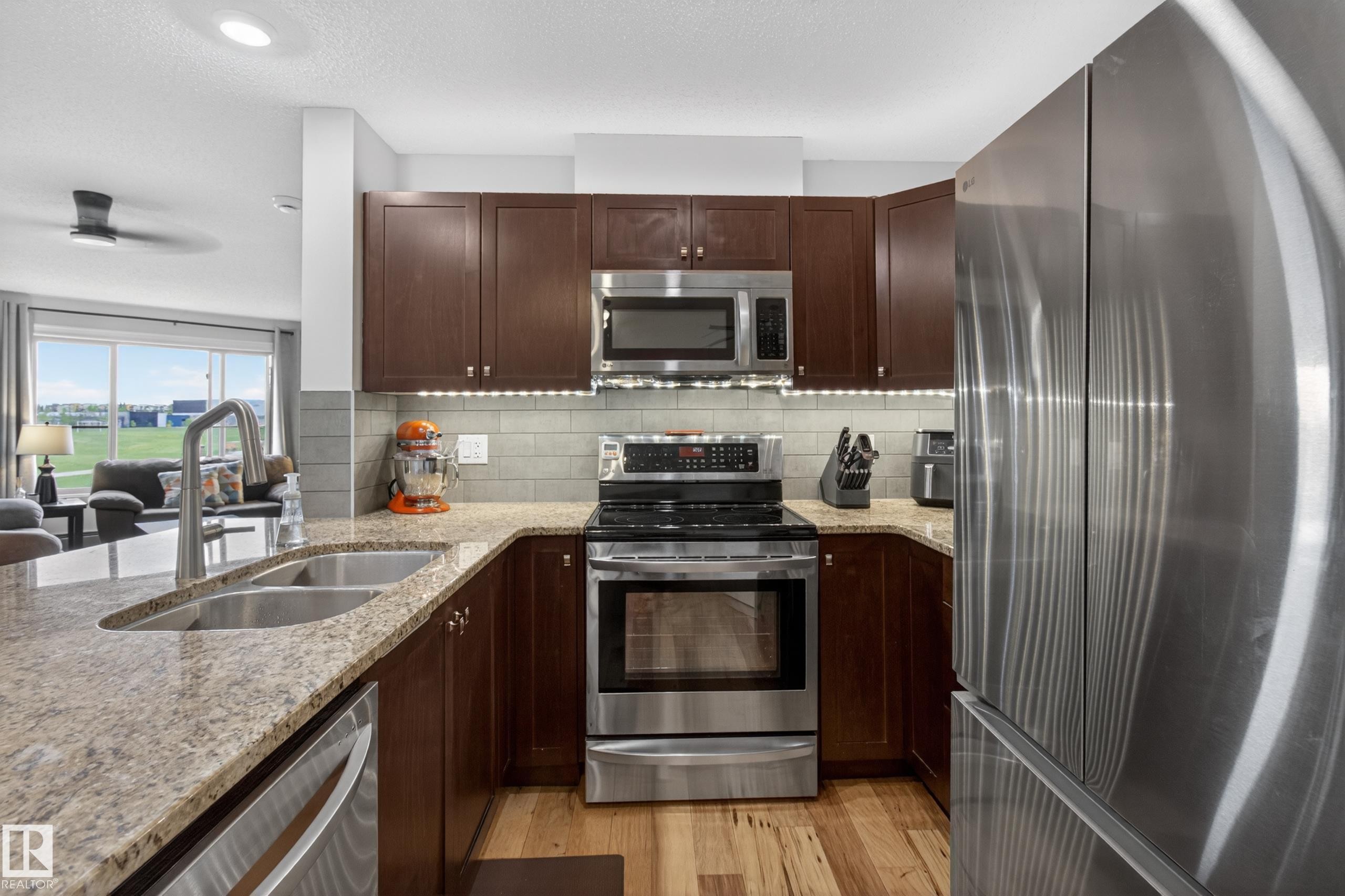 Kitchen featuring rich wood cabinetry, stainless steel appliances, granite countertops, a double basin sink with a high-arc faucet, and hardwood flooring - 211 3719 Whitelaw Lane, Edmonton, AB