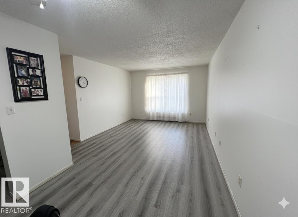 Spacious room featuring light gray wood-finish flooring, white walls, and a window with sheer white curtains - 212 2620 Mill Woods Road E, Edmonton, AB