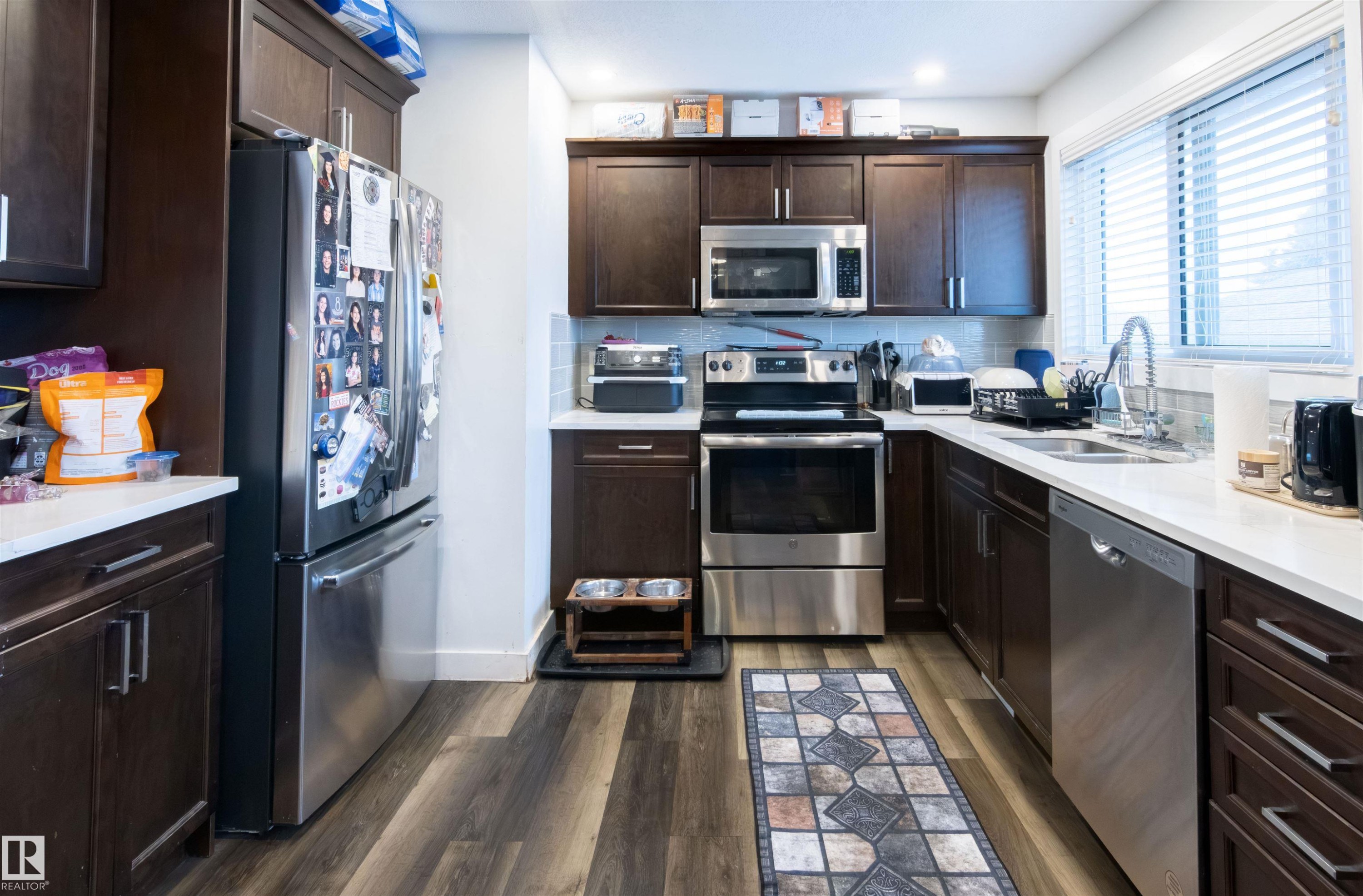 Kitchen featuring wood-finish flooring, dark wood cabinetry, white countertops, a mosaic tile backsplash, and stainless steel appliances including a refrigerator, range, microwave, and dishwasher - 4004 88 Street Nw, Edmonton, AB - Indoor Photo Showing Kitchen With Double Sink