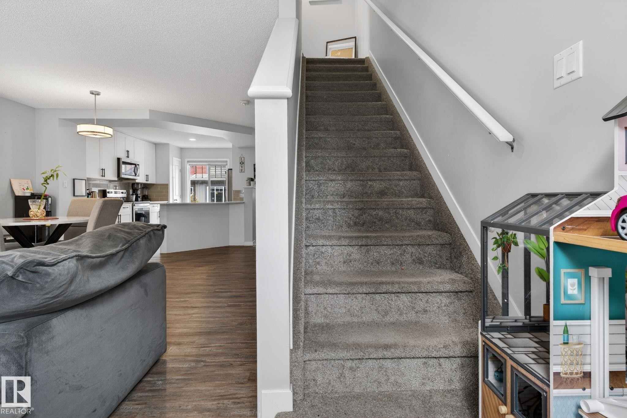 Carpeted staircase with white handrail and baseboards, opening to a living area with wood-finish flooring and a kitchen featuring white cabinetry, stainless steel appliances, and a peninsula with countertop seating - 62 3305 Orchards Link, Edmonton, AB
