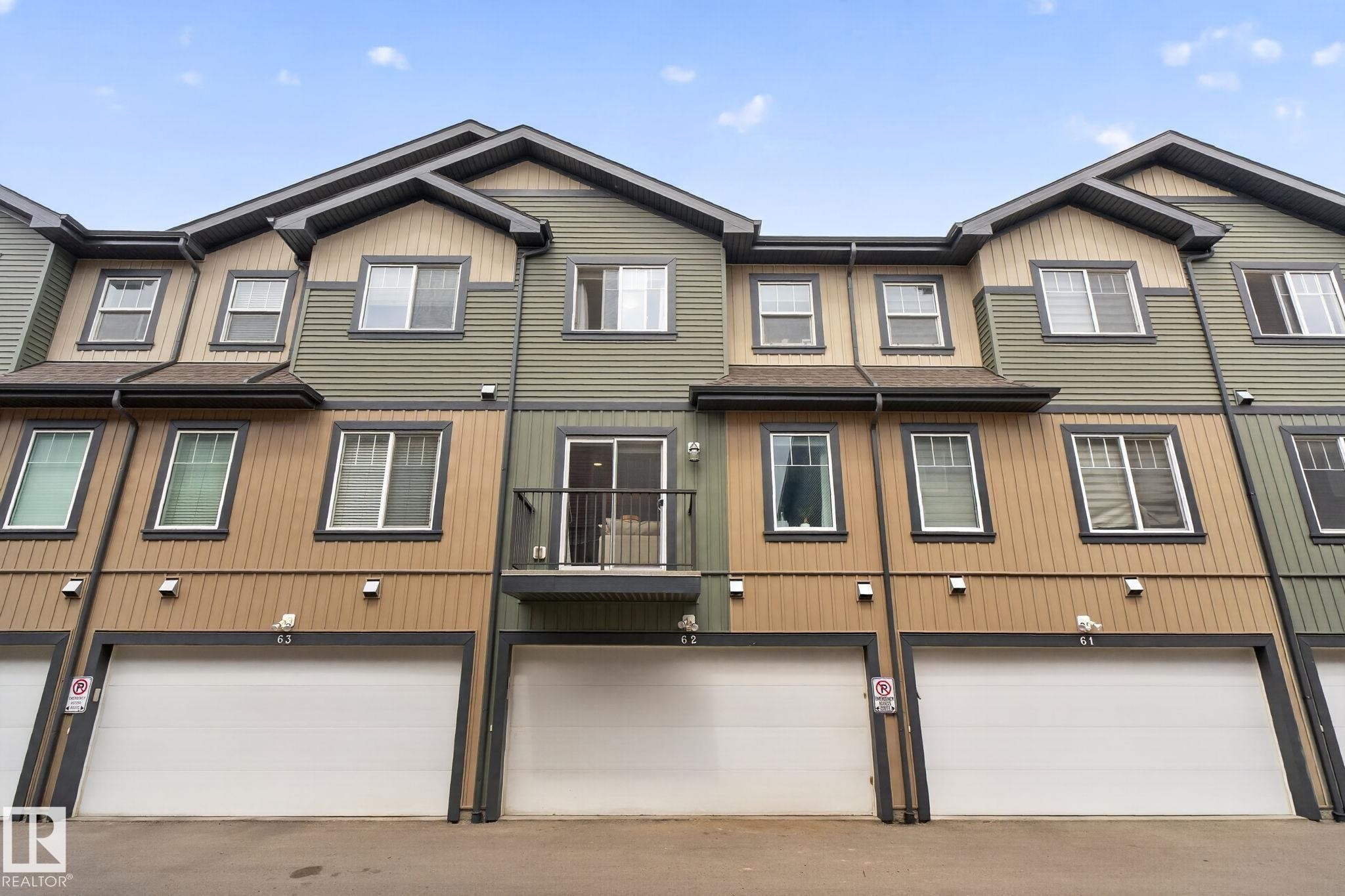 Modern multi-level townhome featuring a mix of horizontal siding and vertical paneling, an integrated single-car garage, a second-story balcony with metal railing, and multiple windows with dark trim - 62 3305 Orchards Link, Edmonton, AB