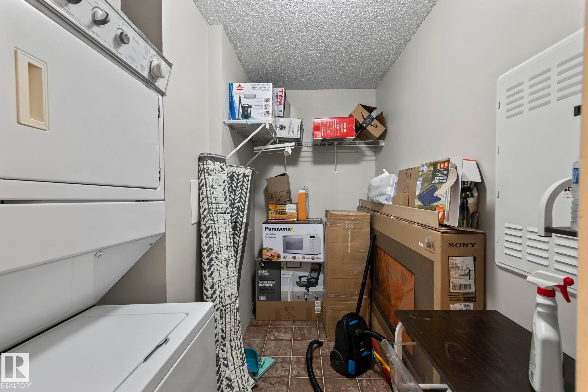 Compact utility area featuring a stacked washer and dryer, tile flooring, and wall-mounted shelving - 2106 9357 Simpson Dr Nw, Edmonton, AB - Indoor Photo Showing Laundry Room