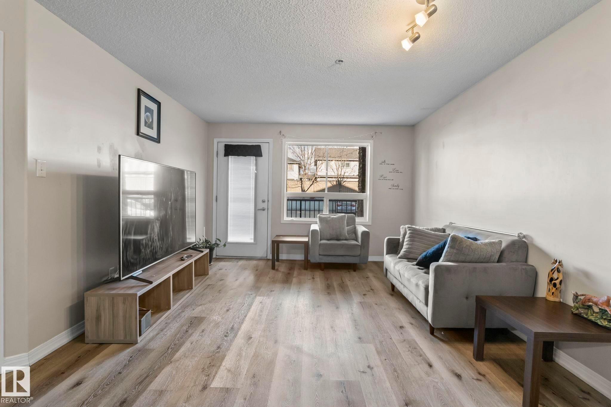 Spacious living area featuring wood-finish flooring, neutral wall tones, a large window, and a white exterior door - 2106 9357 Simpson Dr Nw, Edmonton, AB - Indoor Photo Showing Living Room
