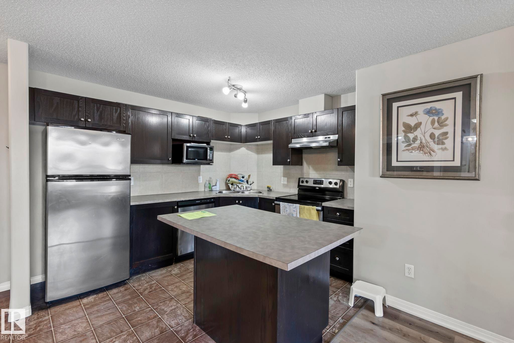 Kitchen featuring dark wood-finish cabinetry, stainless steel appliances, a central island with a grey countertop, and tiled flooring - 2106 9357 Simpson Dr Nw, Edmonton, AB - Indoor Photo Showing Kitchen With Stainless Steel Kitchen