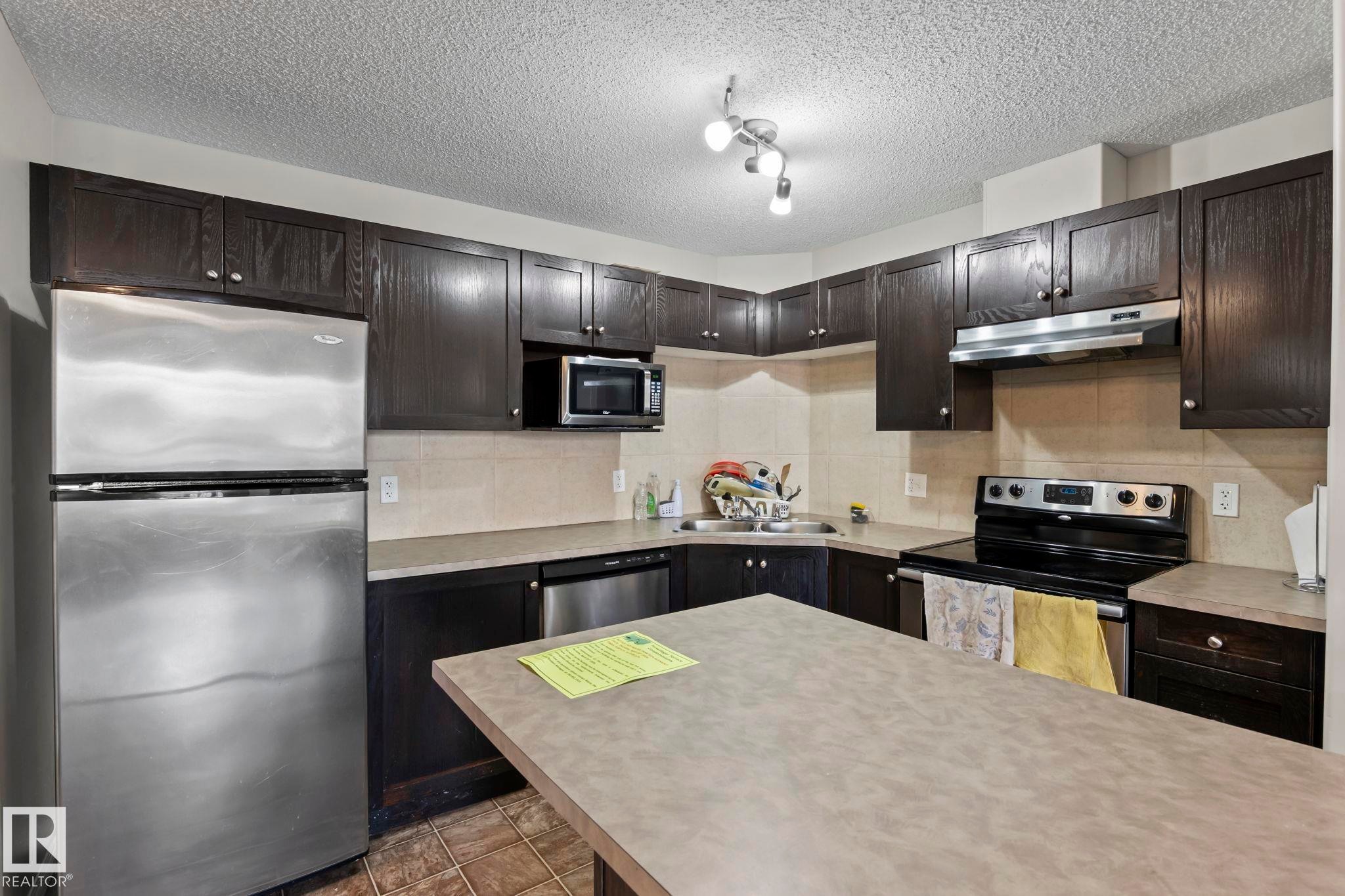 Kitchen featuring dark wood-finish cabinetry, stainless steel appliances, a tile backsplash, and a central island with a solid surface countertop - 2106 9357 Simpson Dr Nw, Edmonton, AB - Indoor Photo Showing Kitchen With Stainless Steel Kitchen With Double Sink With Upgraded Kitchen