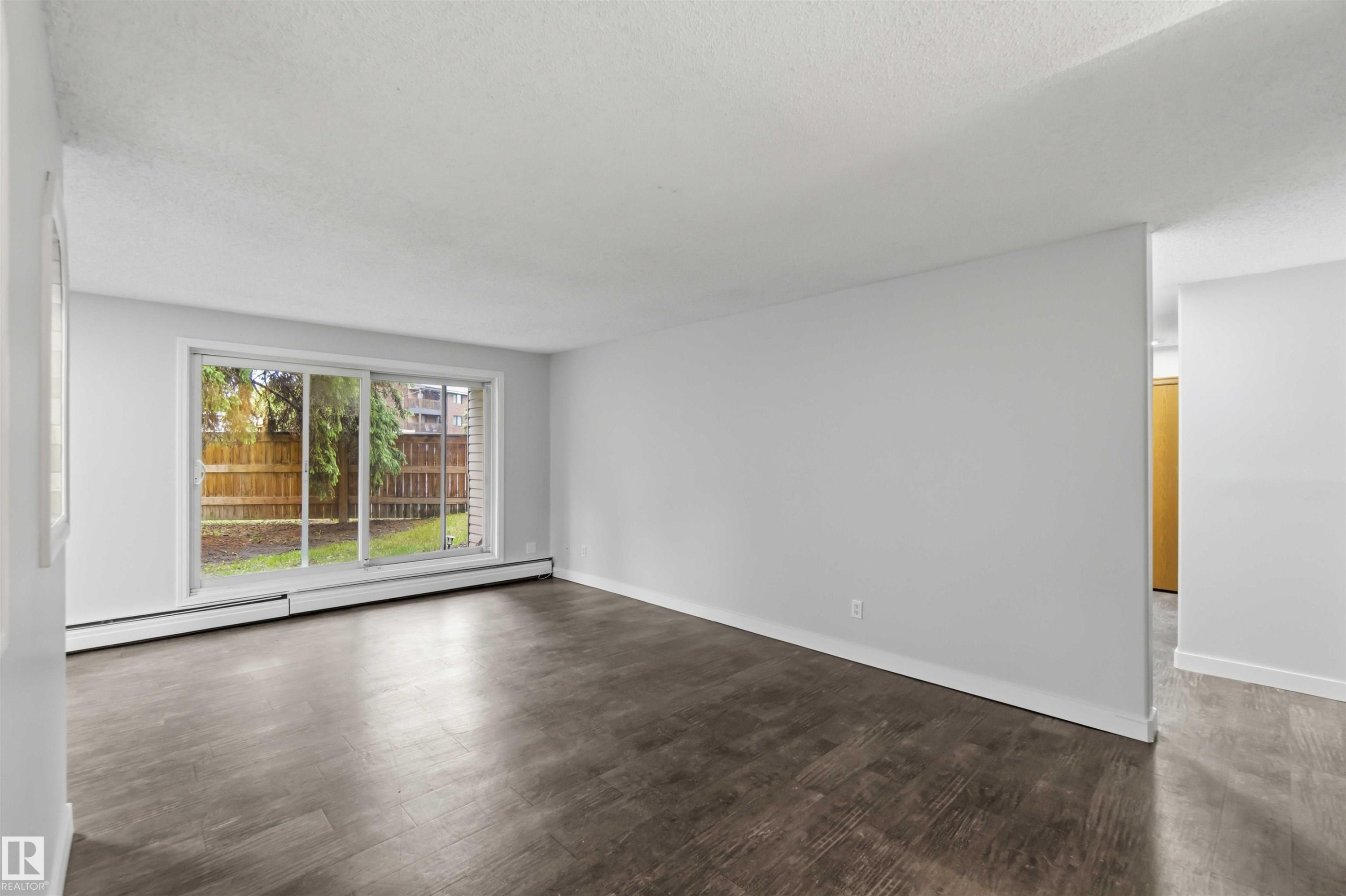 Spacious room featuring wood-finish flooring, a large sliding glass door, white baseboards, and a neutral wall color - #121 5125 Riverbend Rd Nw Nw, Edmonton, AB - Indoor