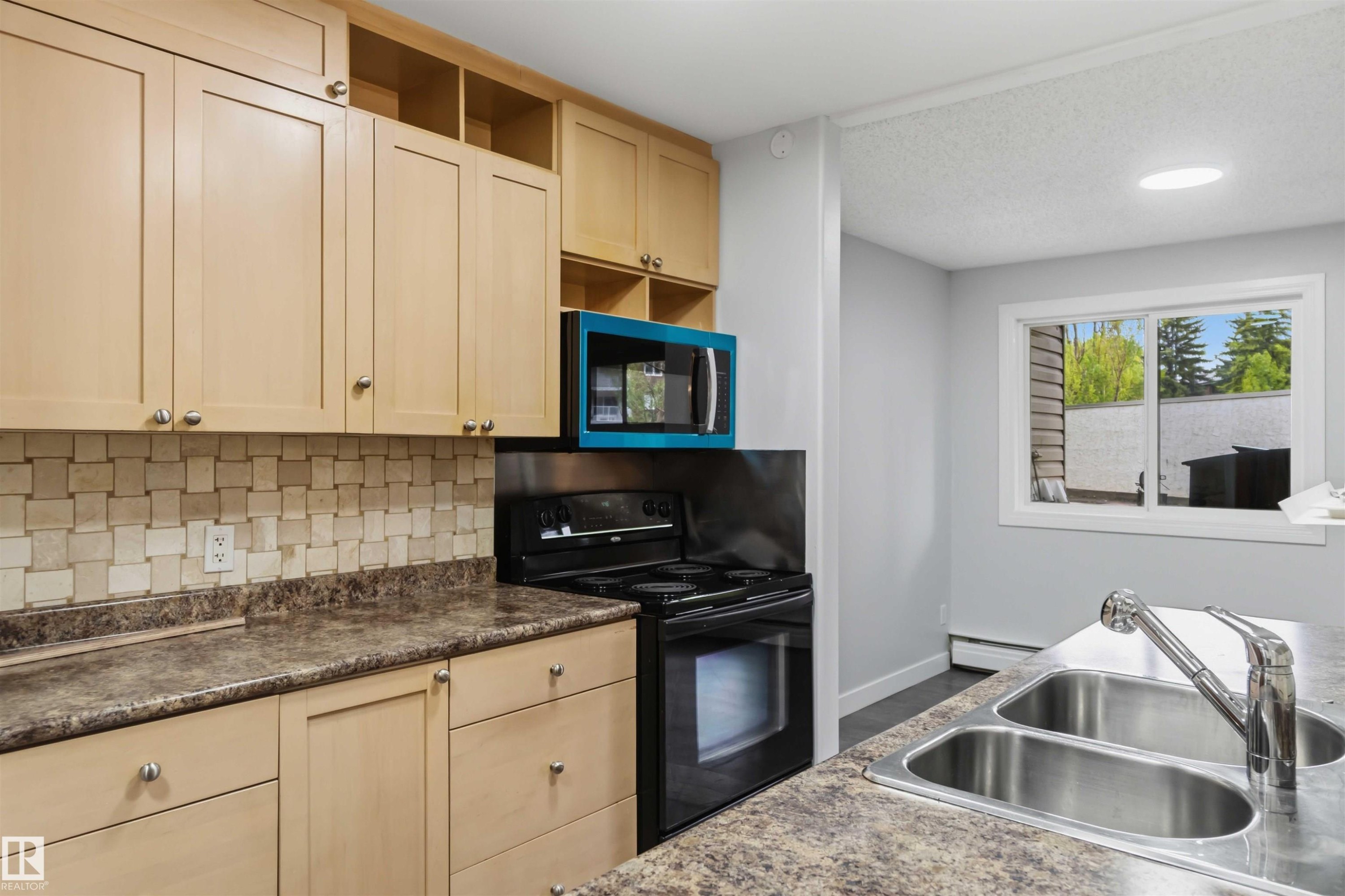 Kitchen featuring light wood-finish cabinetry, patterned tile backsplash, and dark patterned countertops - #121 5125 Riverbend Rd Nw Nw, Edmonton, AB - Indoor Photo Showing Kitchen With Double Sink